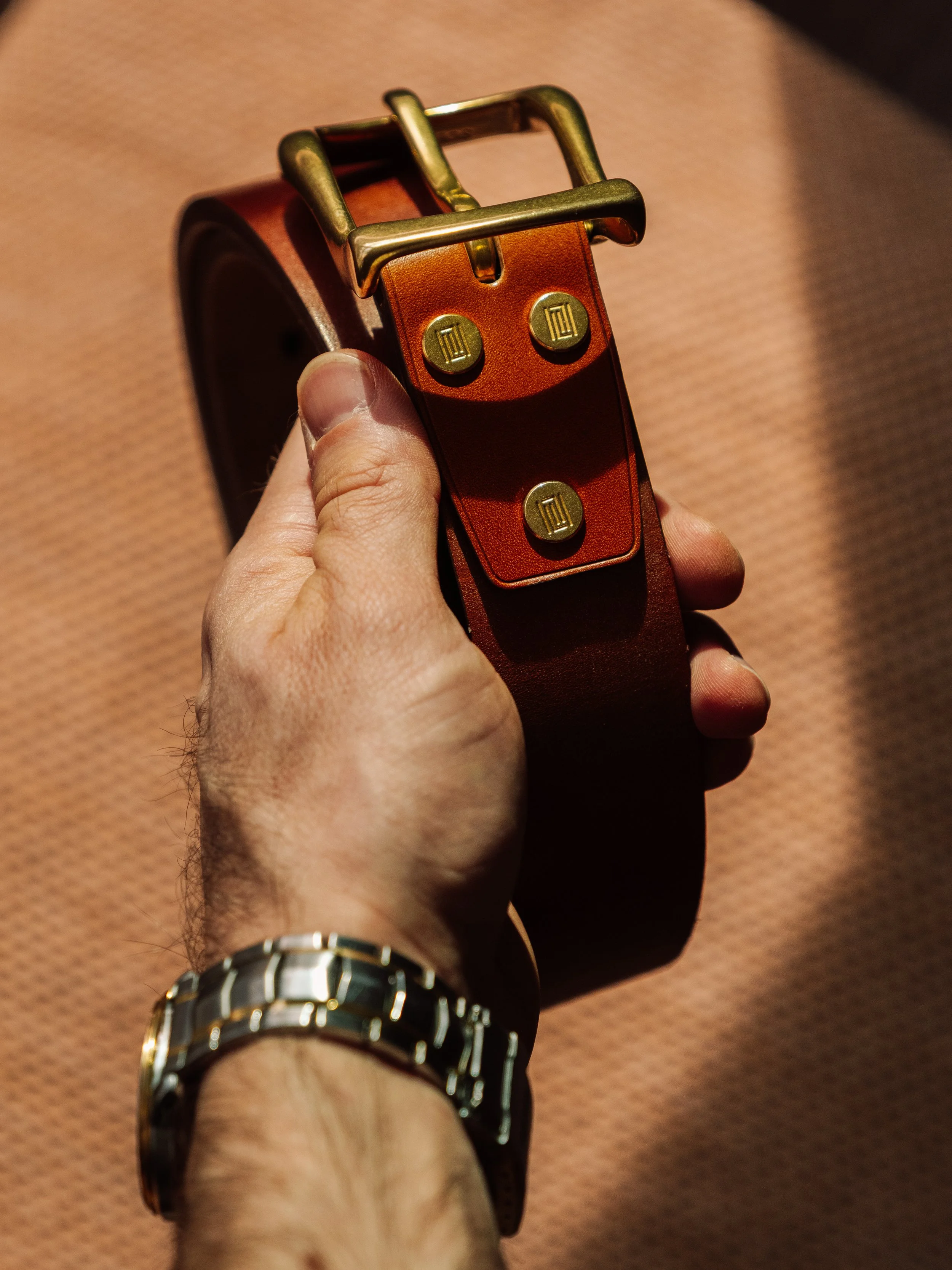 Commercial product photography Campaign. Close-up of a hand holding a brown leather belt with a gold buckle, against a textured background.
