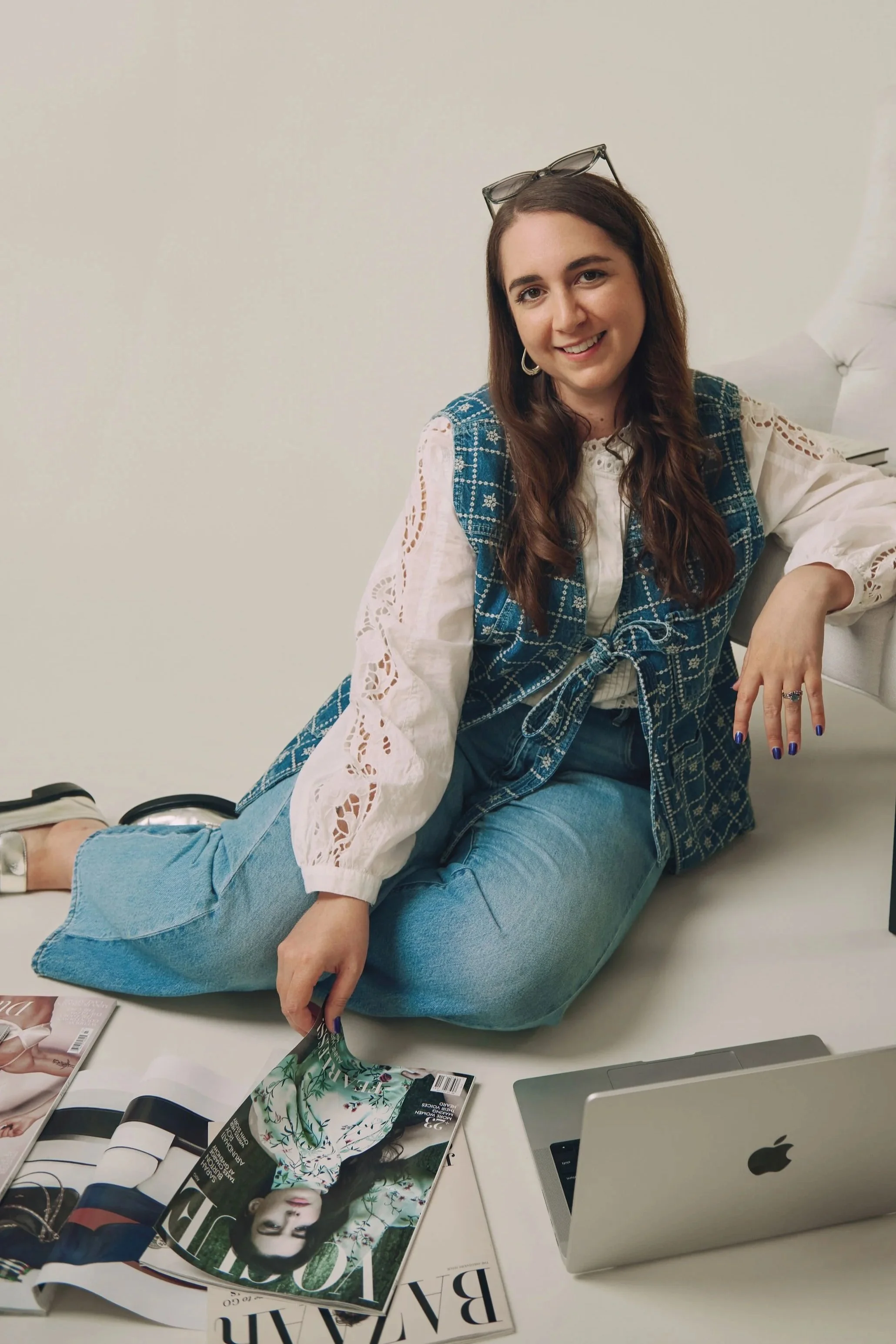 Young woman sitting on a white bed surrounded by magazines and an open laptop, smiling at the camera.