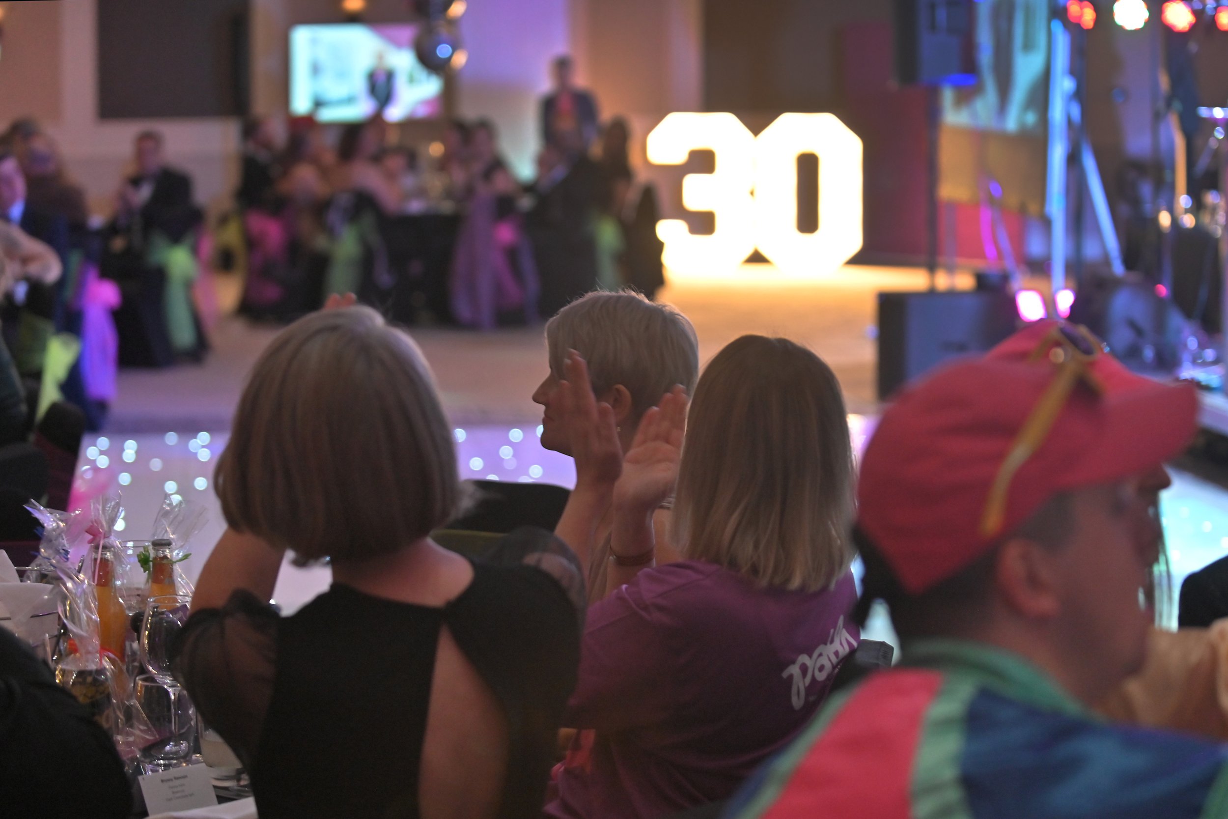 Musicians performing on stage, with one jumping mid-air playing an electric guitar, and others playing bass and drums, in a dimly lit venue.