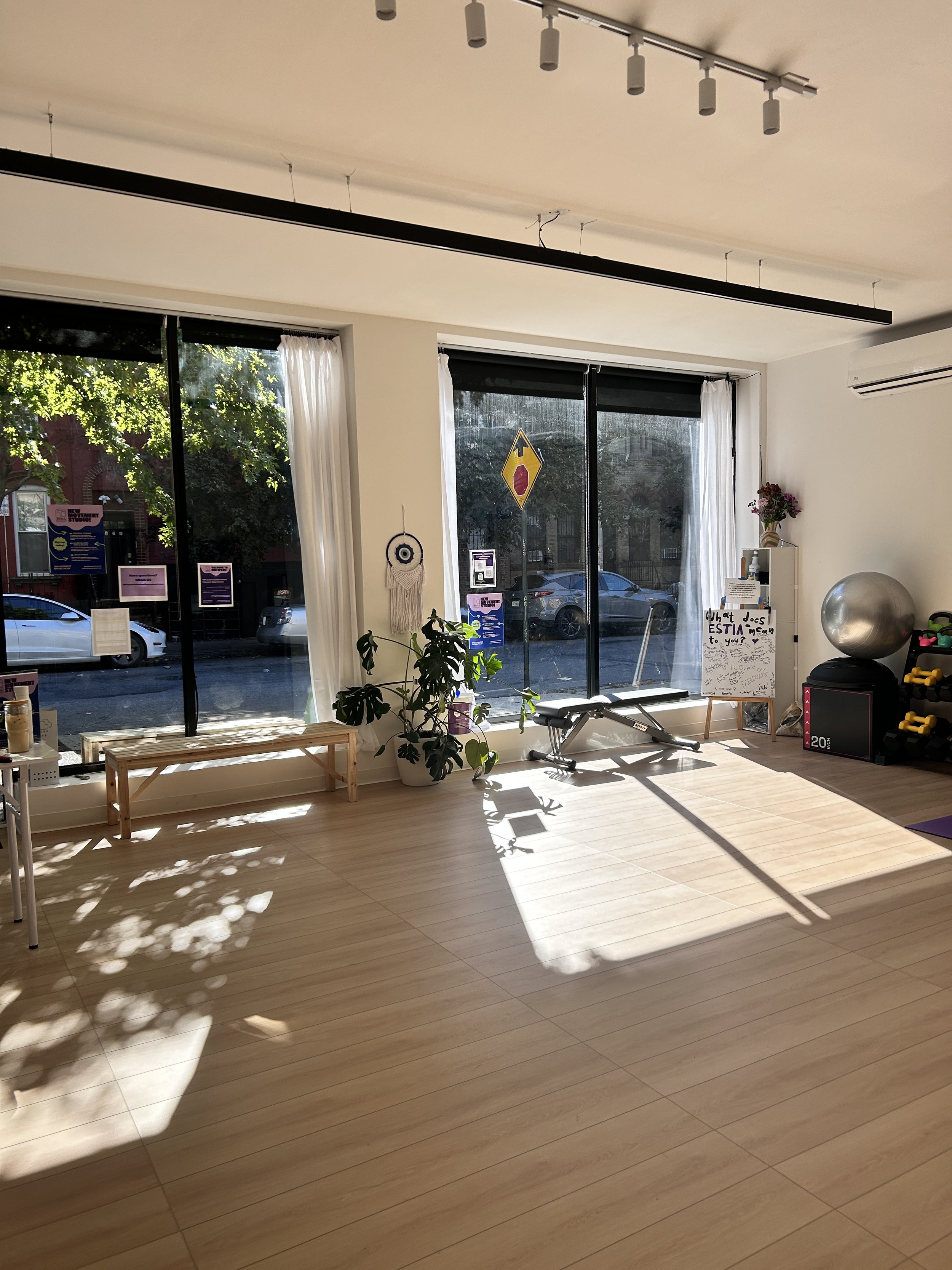 Open studio space at ESTIA House of Movement featuring fitness equipment, seating, large front windows, and natural light.