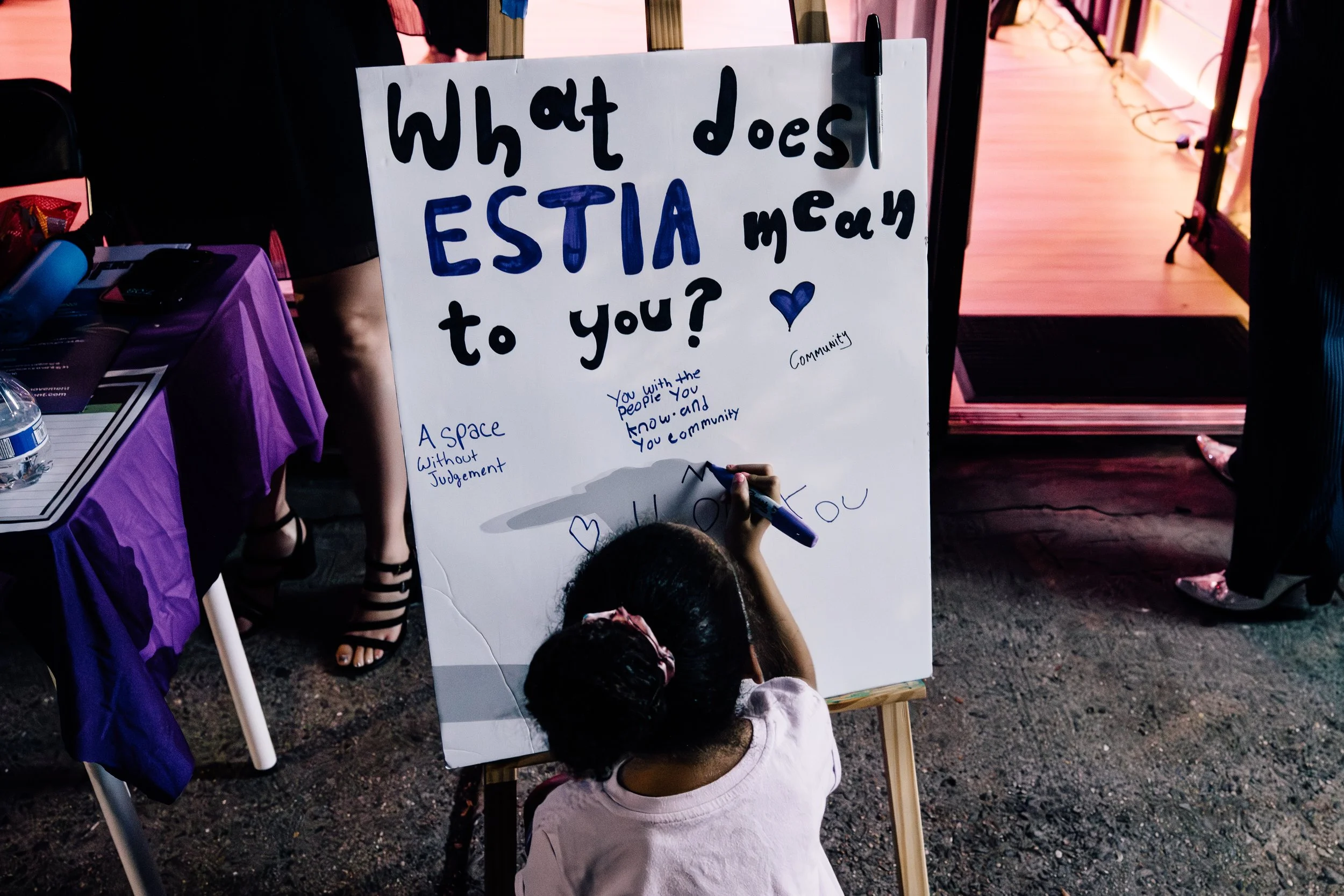 Child writing on a community message board that reads, "What does ESTIA mean to you?" with handwritten responses from community members.