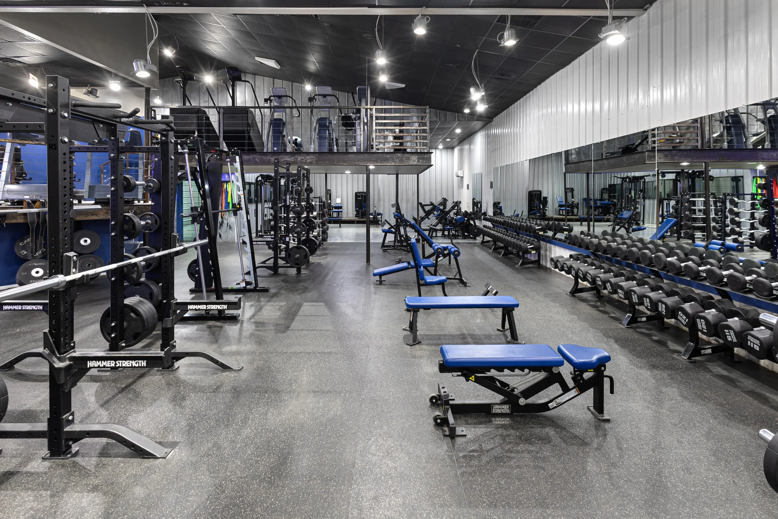 Empty gym with various weightlifting equipment, benches, and free weights, mirrored walls, black ceiling, and bright overhead lights.