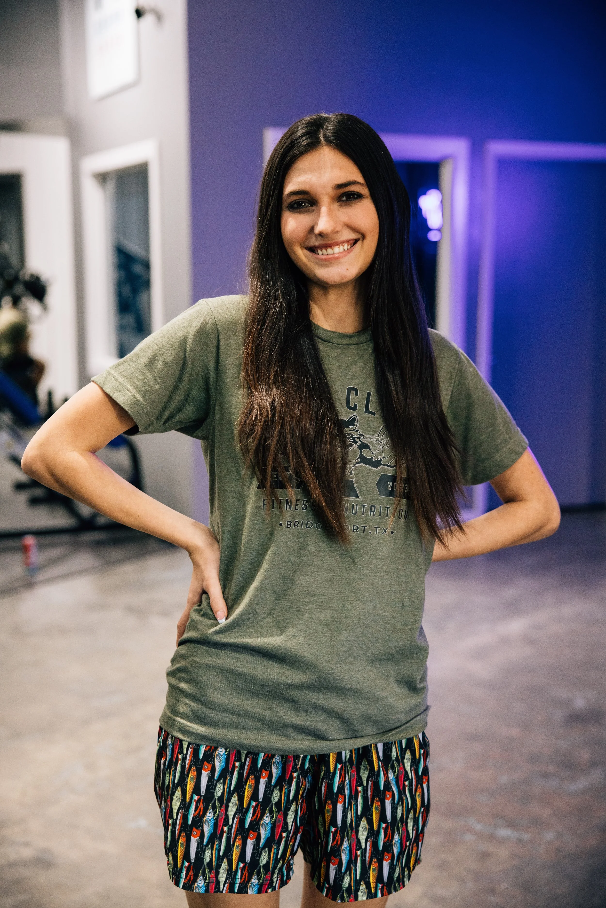 Smiling young woman with long dark hair, wearing a graphic t-shirt and colorful shorts, standing with hands on hips inside a gym or fitness center.