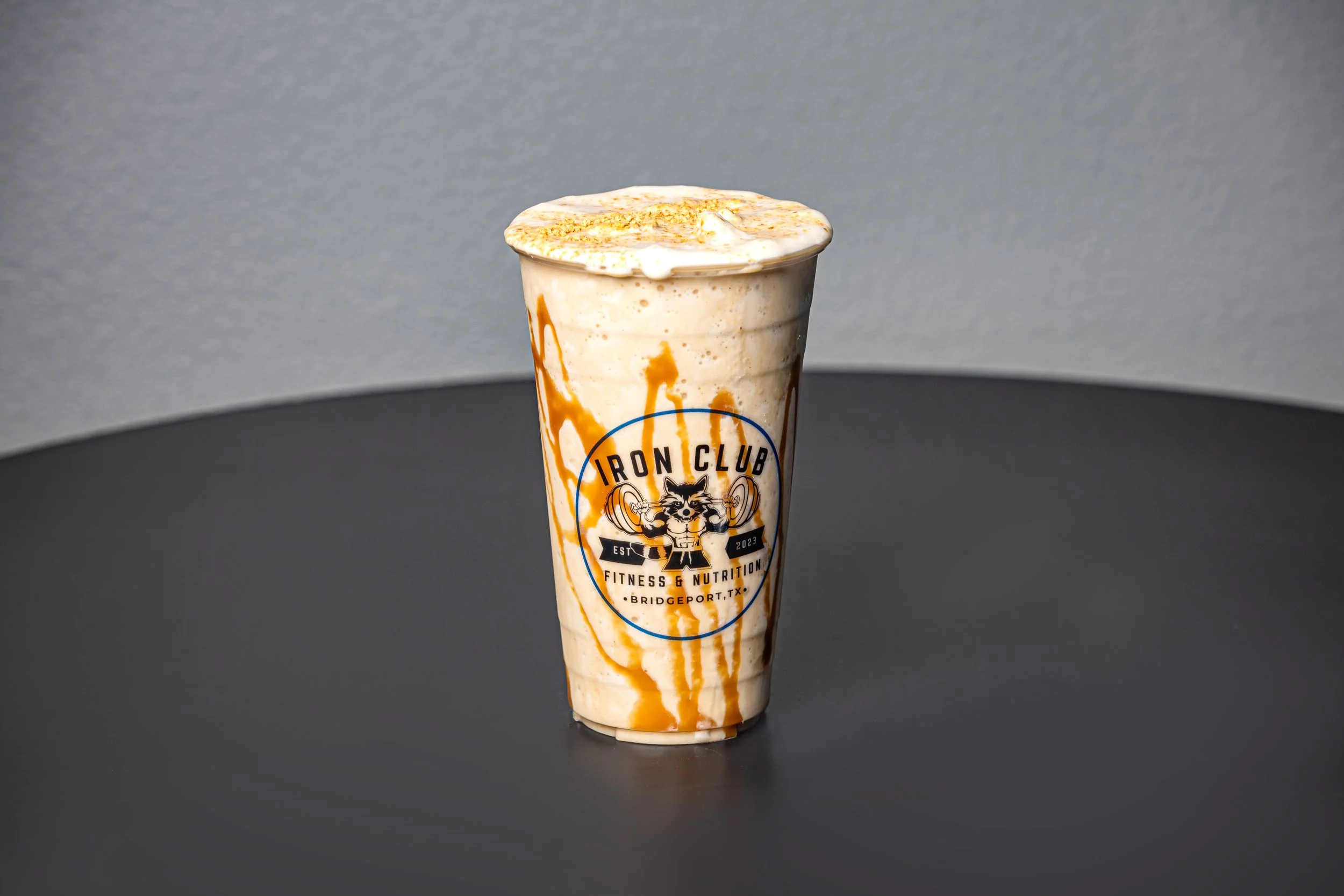 A caramel frappuccino with whipped cream and caramel drizzle in a clear plastic cup with the Iron Club logo, on a black surface against a gray background.