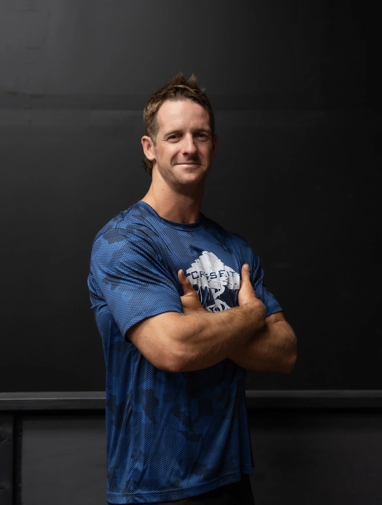 A young man with light brown hair, smiling with arms crossed, standing against a dark background, wearing a black t-shirt with a graphic design.