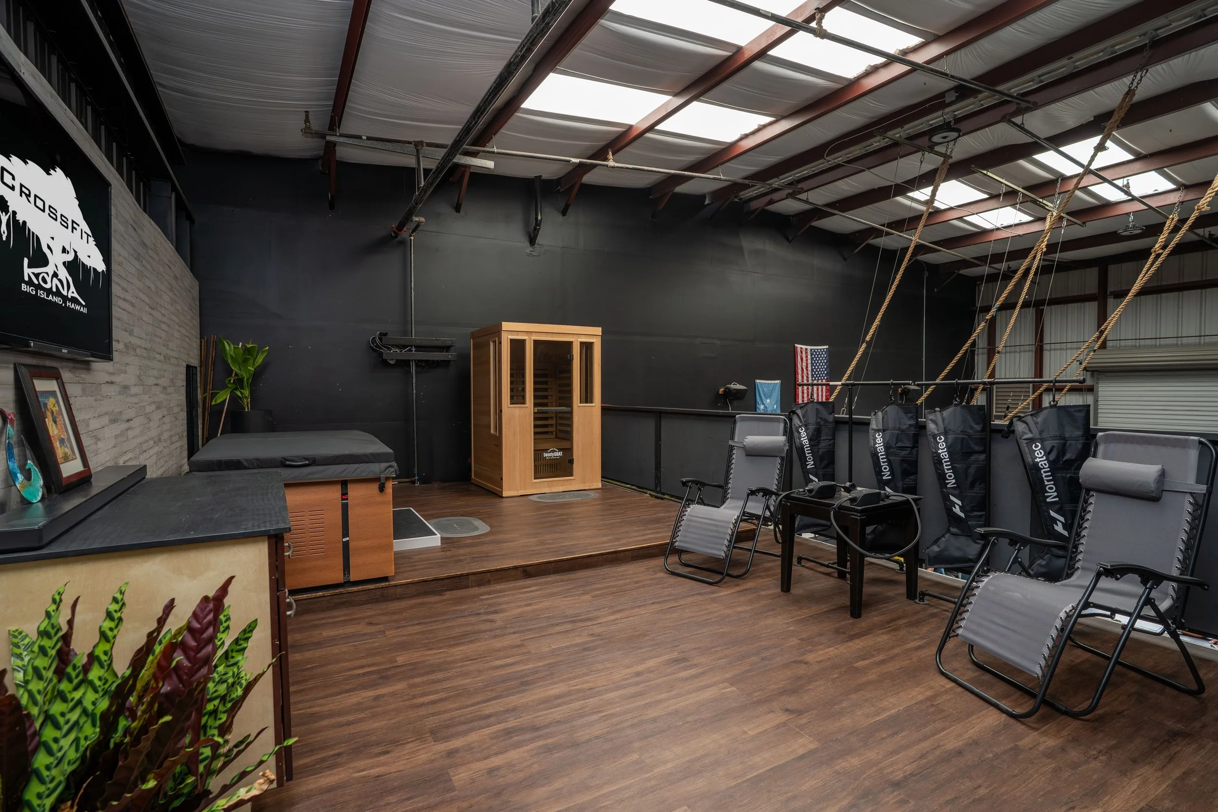 A CrossFit gym with wooden flooring, black and gray walls, and a small raised platform with a sauna in the corner. Ropes hang from the ceiling on the right side, and chairs are arranged along the ramp wall, with American flags and gym equipment visib