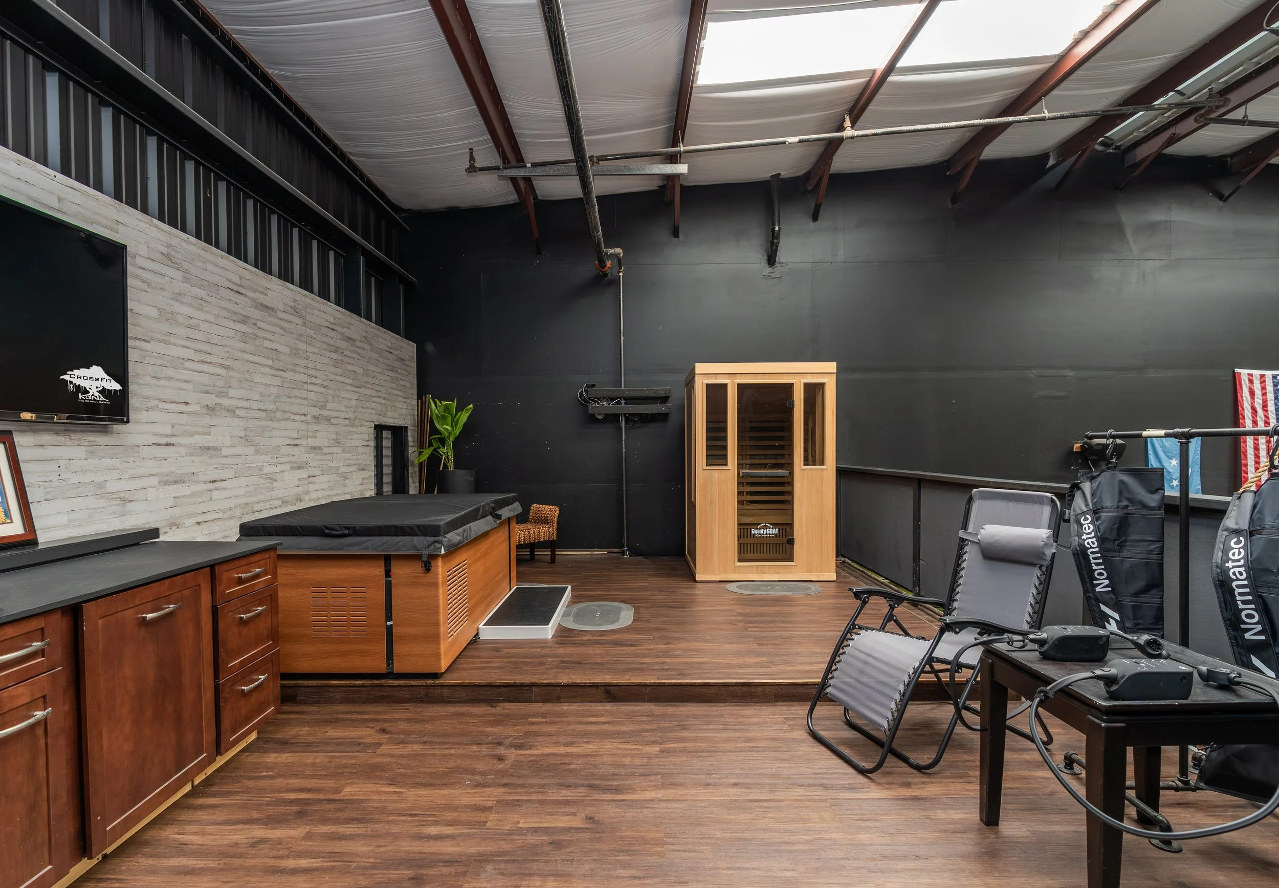 Interior view of a fitness or wellness studio with a sauna, workout equipment, a padded therapy table, a large TV, and wooden and black accents.