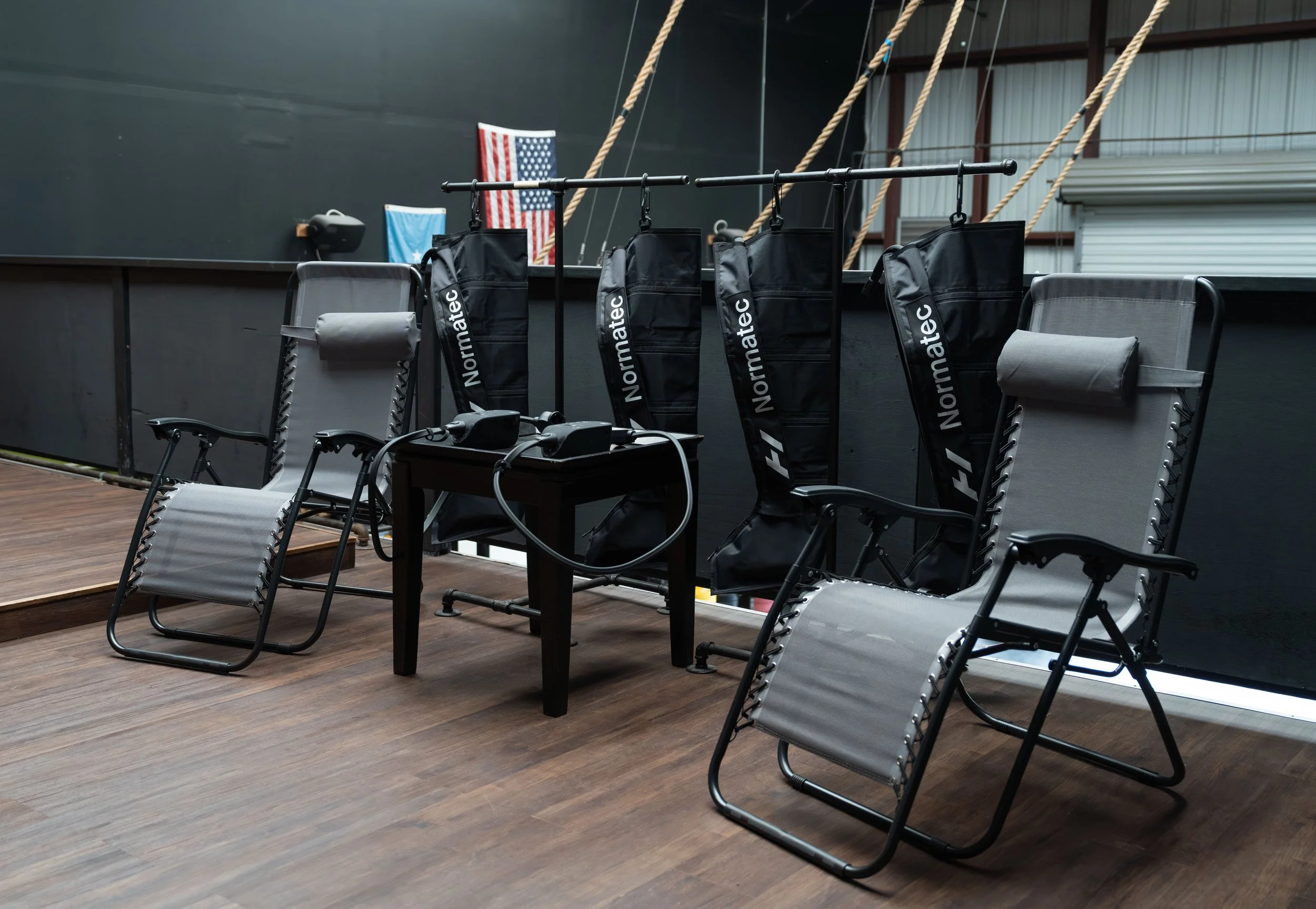 Two gray reclining chairs with black armrests and back support, a black table with a racing simulator steering wheel and pedals, and four black Normatec compression therapy boots hanging on a black rack behind the chairs, in a gym or training facilit