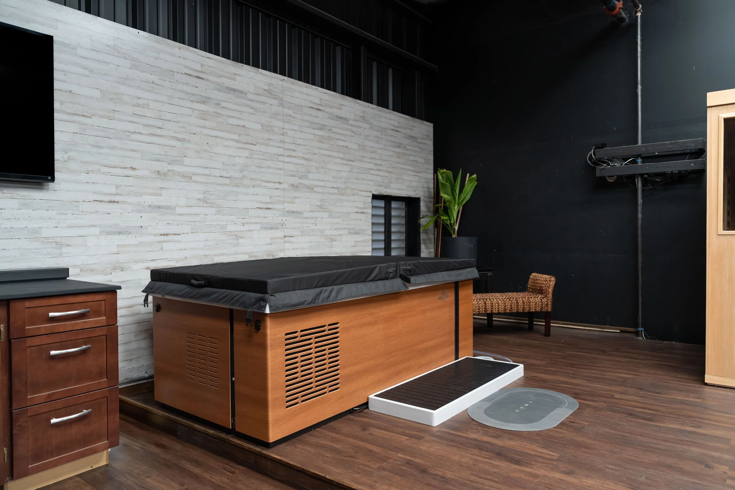 A room with a hot tub, a wooden cabinet, a potted plant, a wicker chair, a large black wall, and a mounted television.