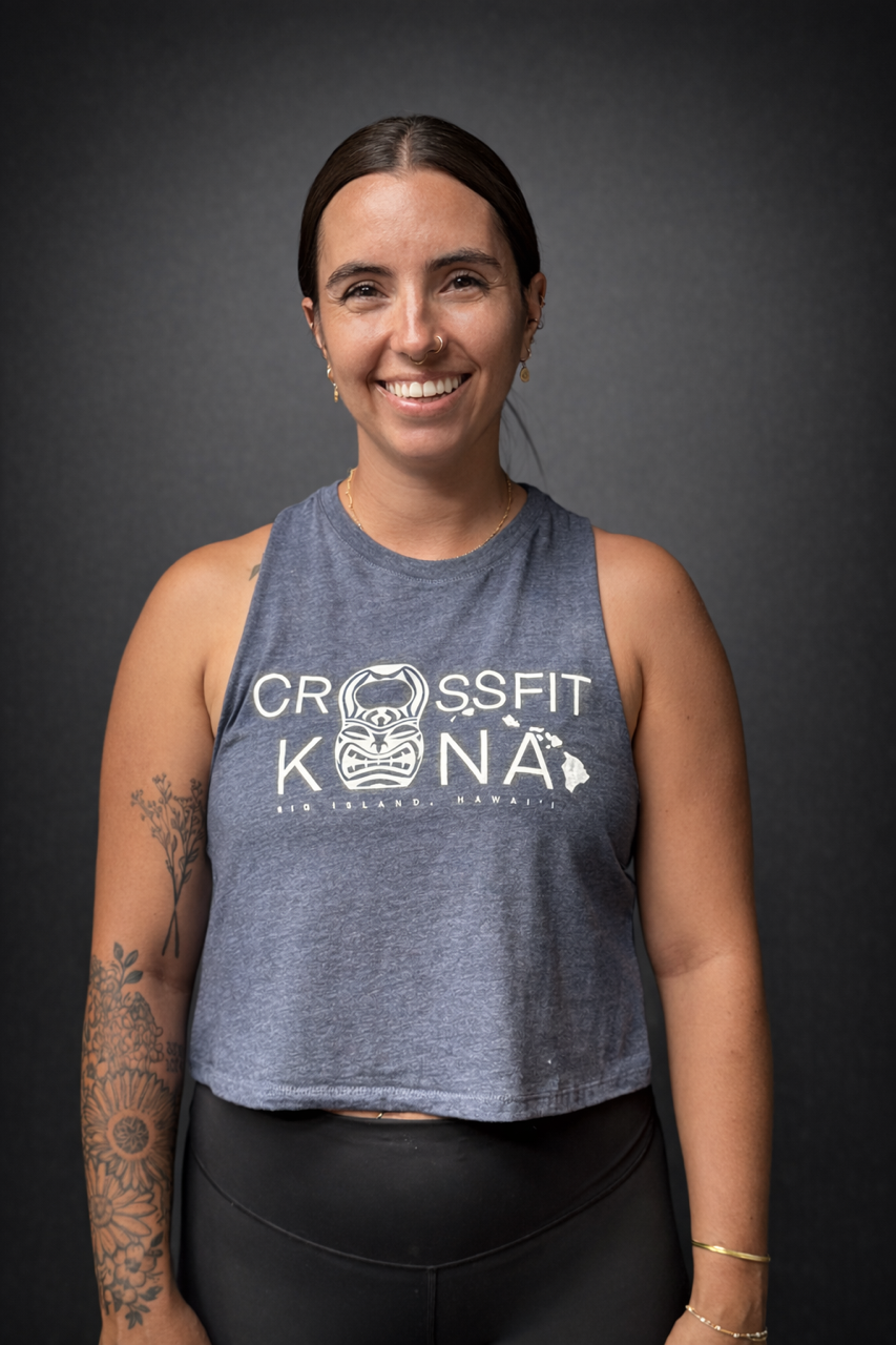 Smiling woman with dark hair, tattoos on her left arm, wearing a pink sleeveless tank top with CrossFit Kona logo, standing against a black background.