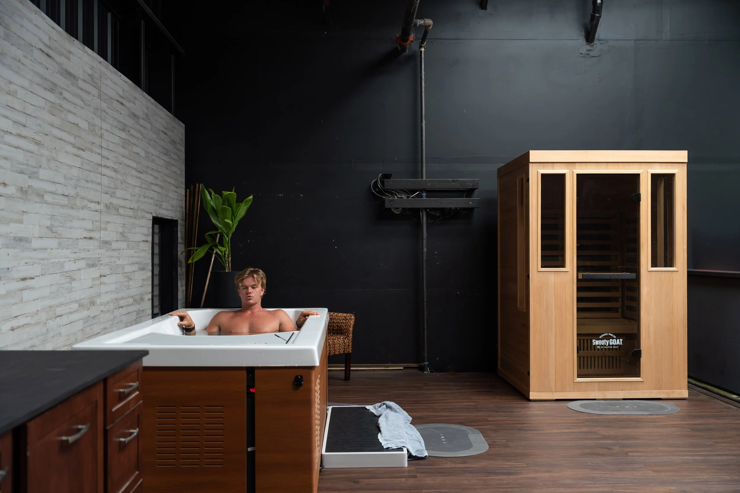 A young man relaxing in a hot tub next to a wooden sauna with glass door in a modern wellness room with dark walls and wooden floors.