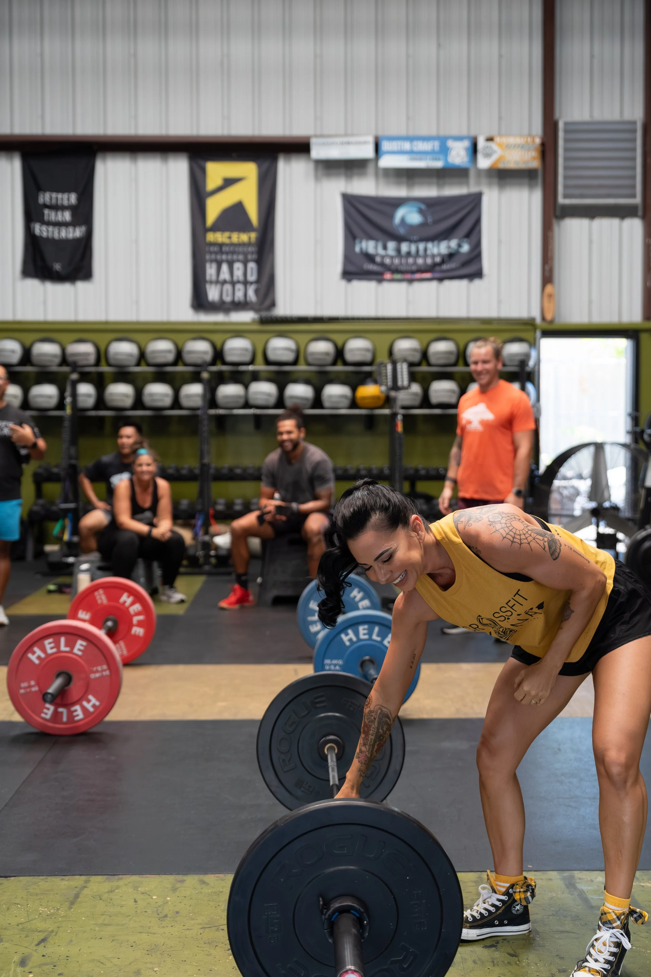 A woman with tattoos smiling as she picks up a barbell in a gym, with people sitting and standing in the background.