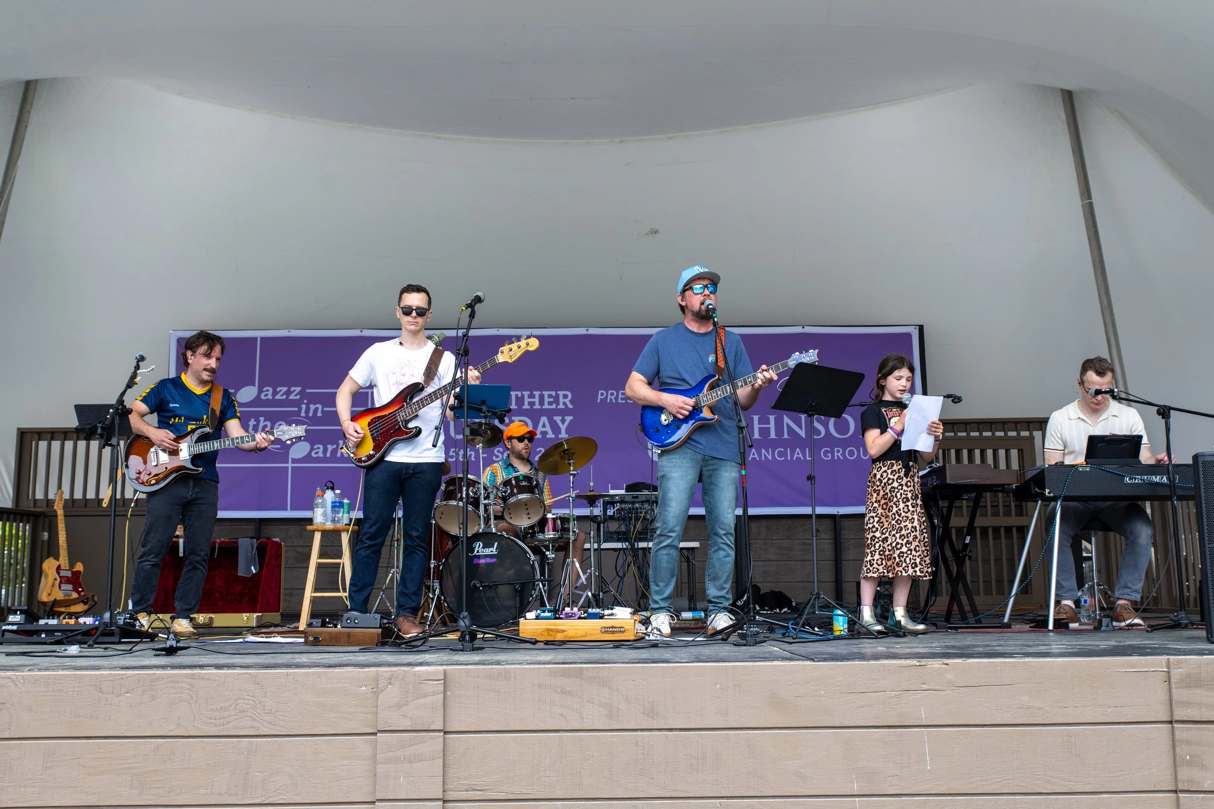 A six-member band performing on an outdoor stage with instruments including guitars, drums, keyboard, and microphone. Four men, one woman, and a young girl are playing music. The backdrop is purple with text.
