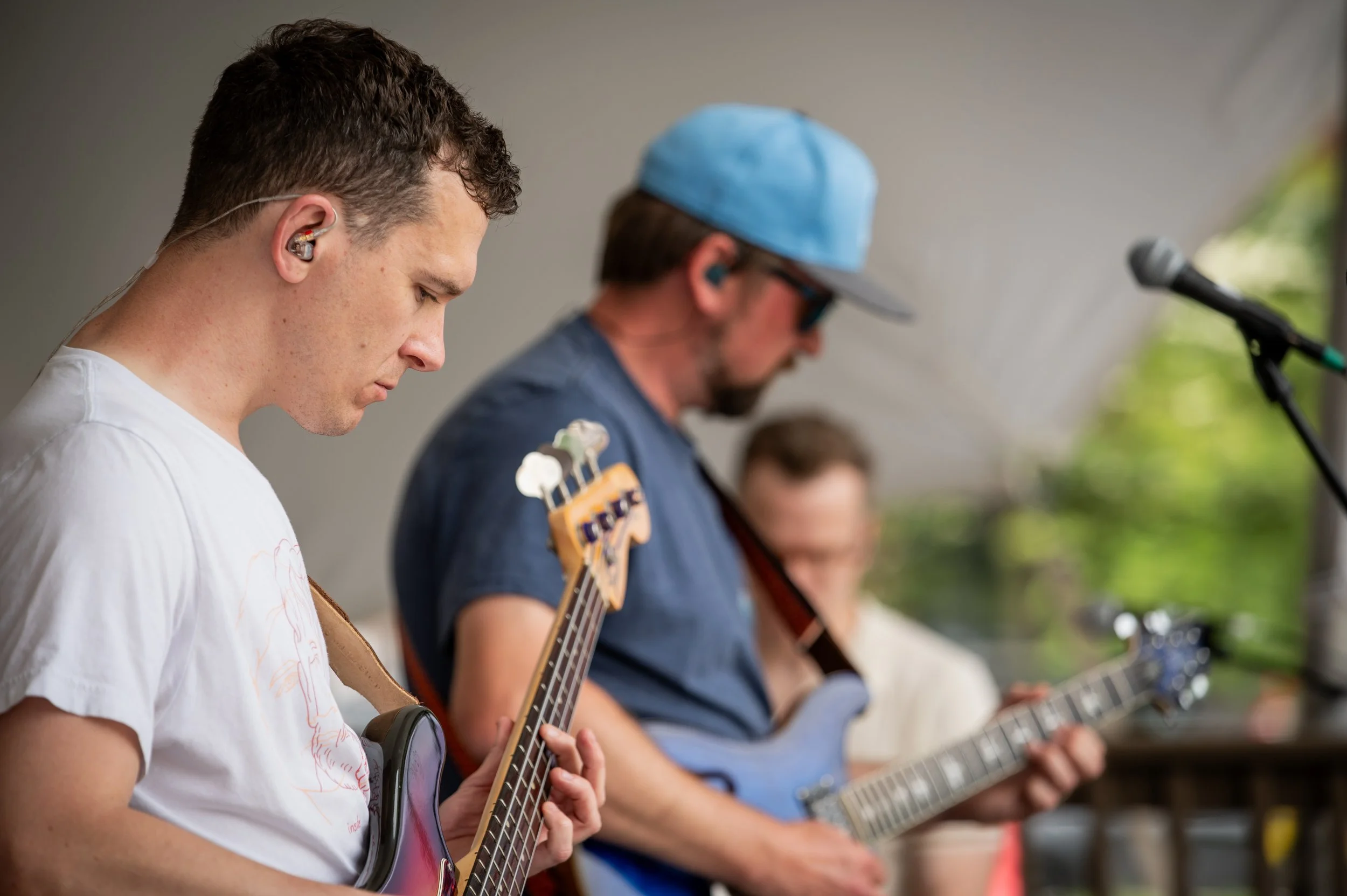 Three male musicians playing guitars and singing at an outdoor event, with a microphone in the foreground.