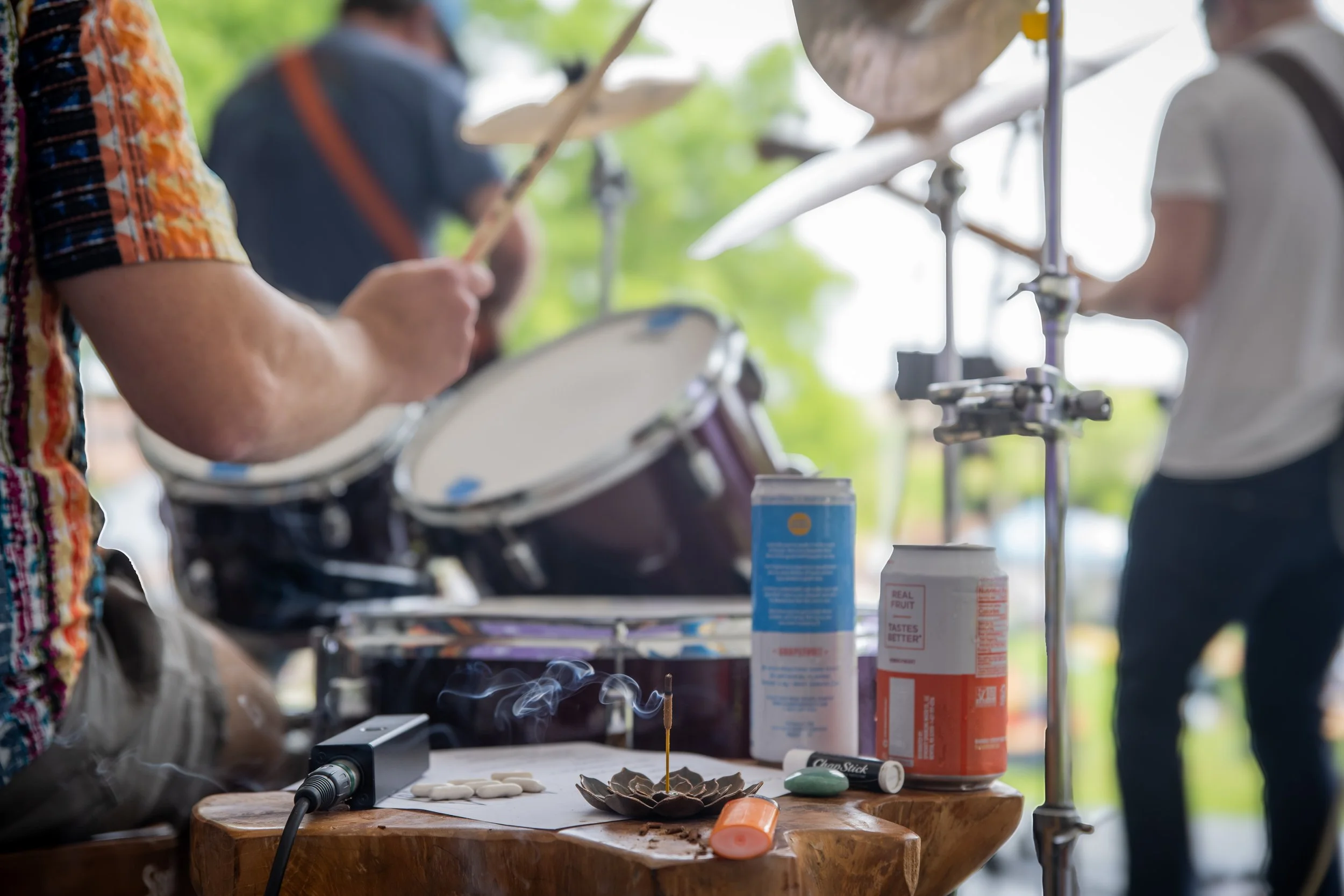 Close-up of a wooden table with smoking incense and medicine, with percussion instruments and people playing drums in the background at an outdoor gathering.