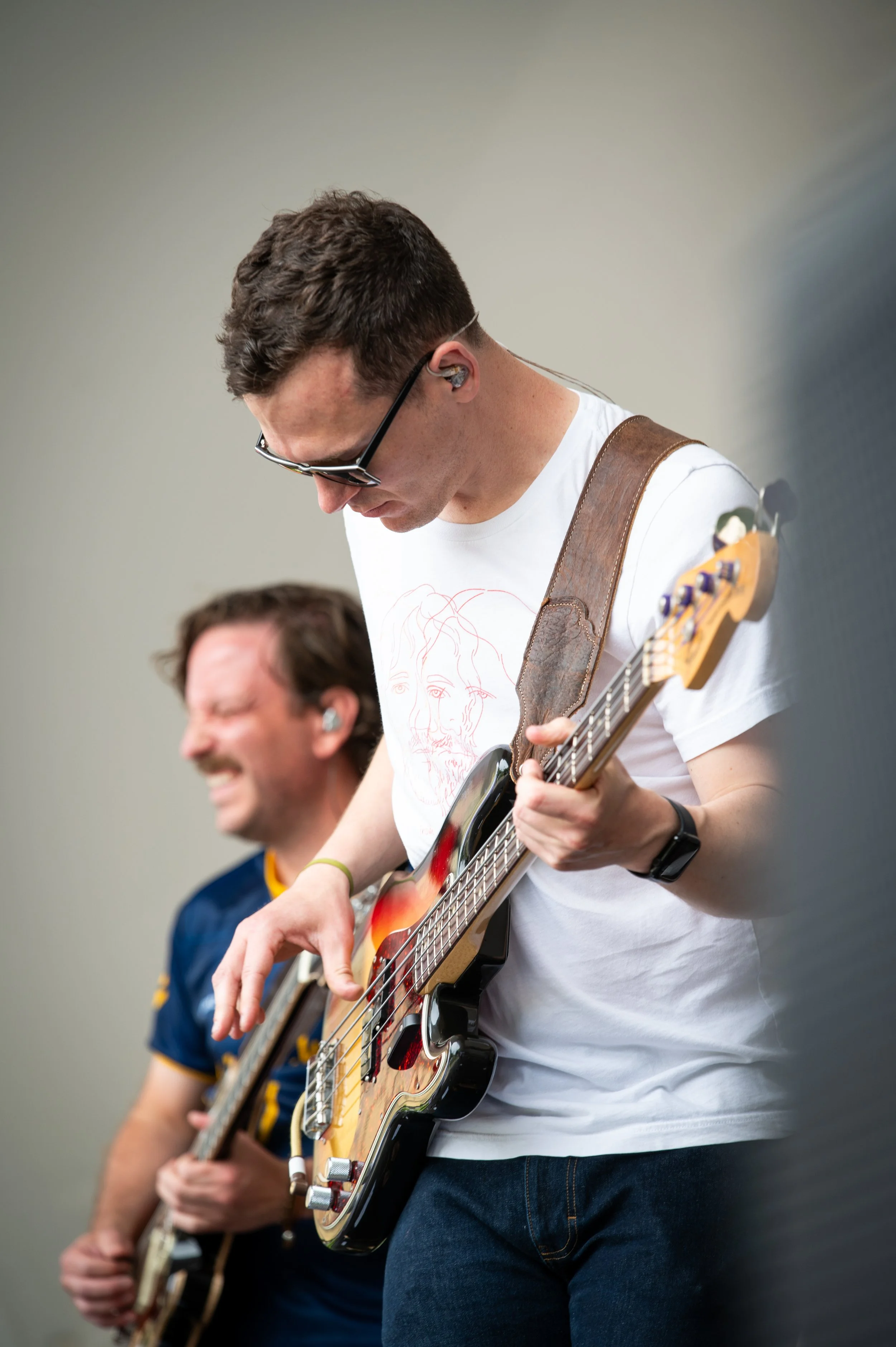 Two young men playing electric guitars, one in focus and the other blurred in the background, both wearing casual t-shirts and in a music practice room.