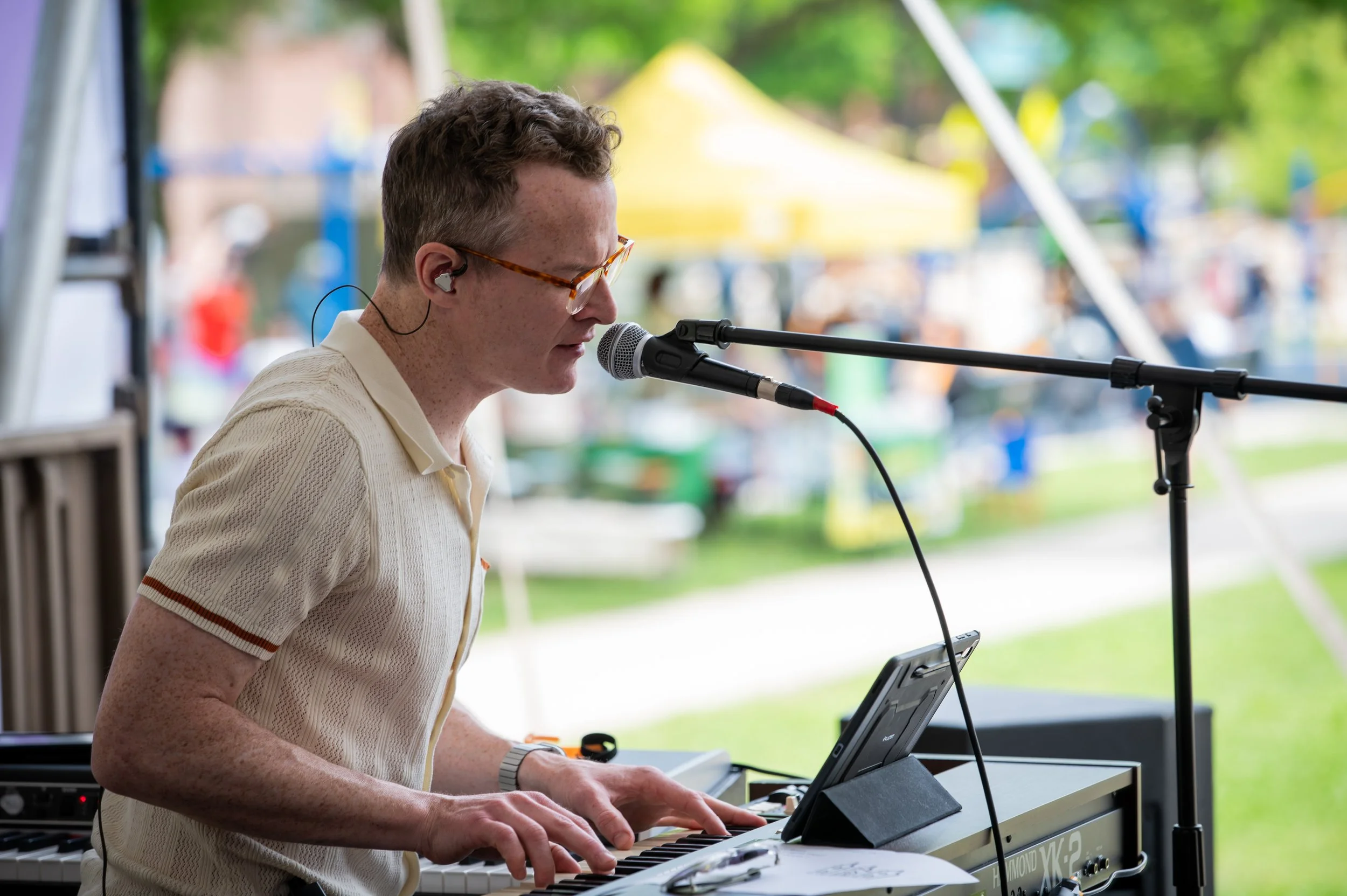 A man with glasses and earphones playing a keyboard and singing into a microphone at an outdoor event.