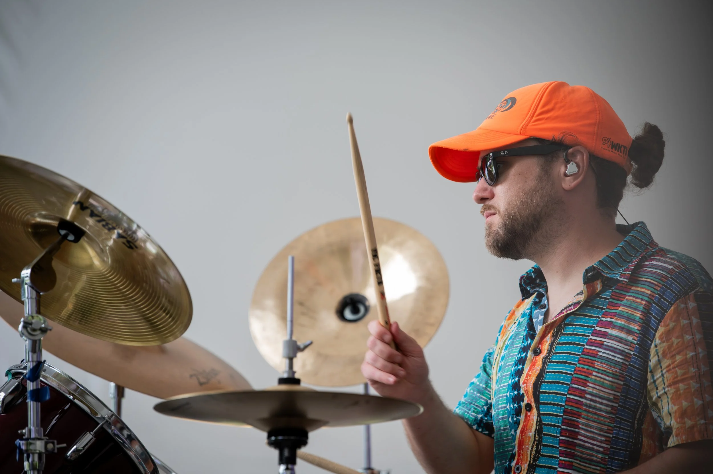 A man wearing sunglasses, an orange cap, and a colorful short-sleeve shirt playing drums with a focused expression.