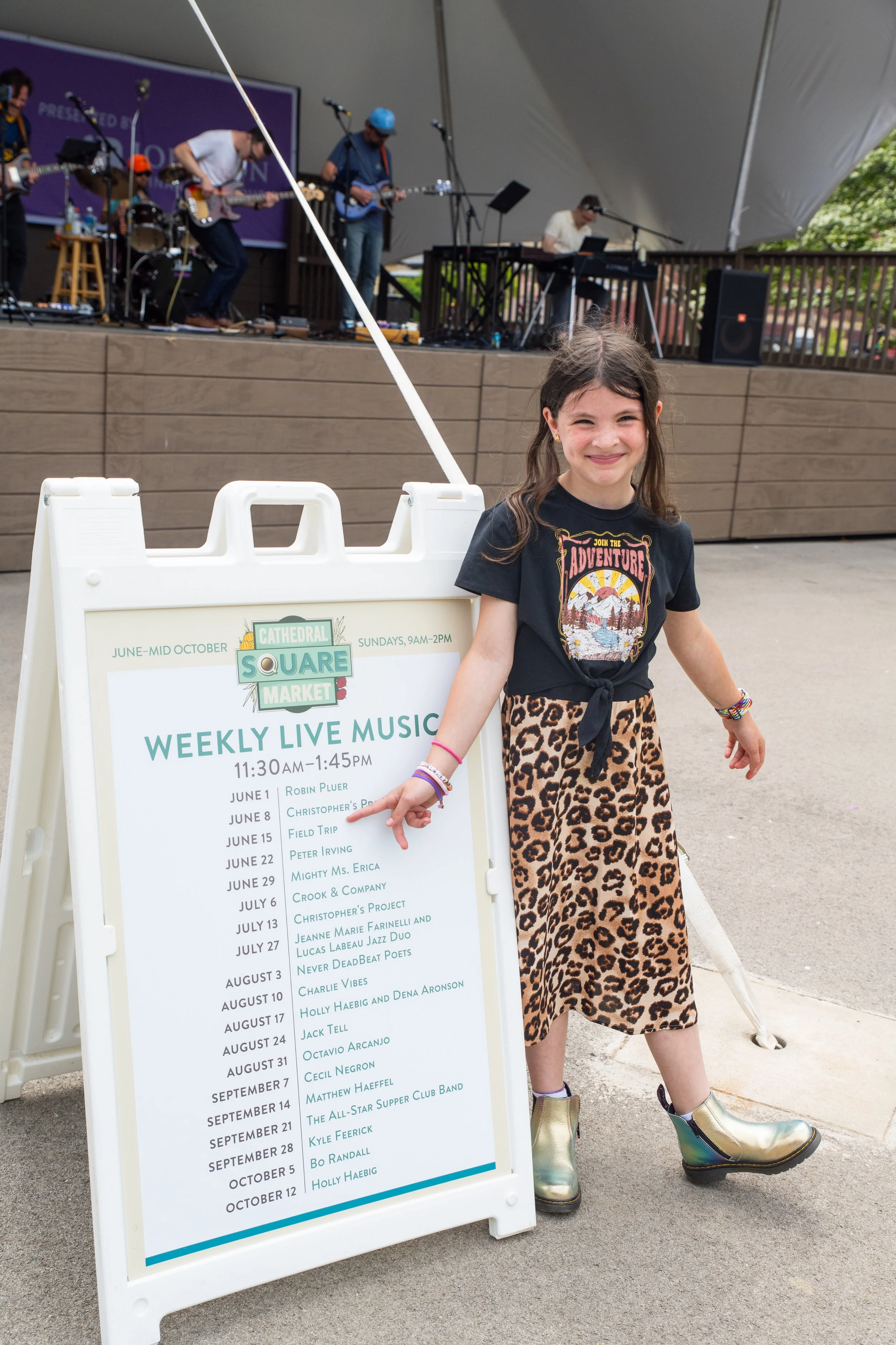 A young girl standing next to a signboard with a weekly live music schedule at the Cathedral Square Market, with a band performing on stage in the background.