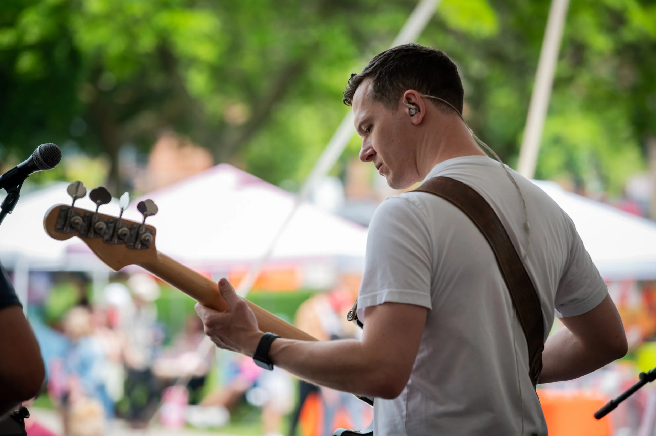 Man playing electric guitar at an outdoor event with trees and tents in the background.