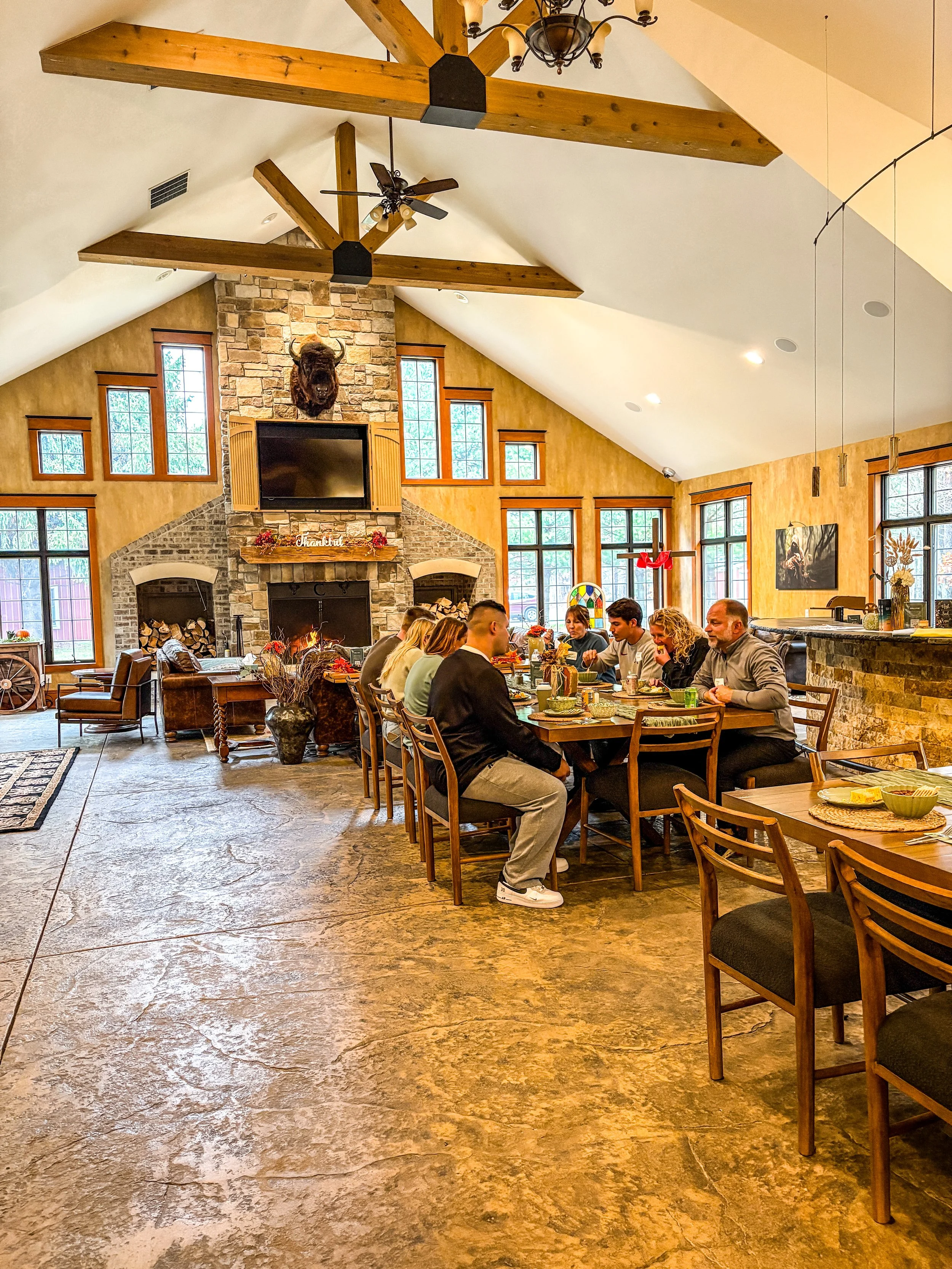 Spacious gathering place with a 18' vaulted ceiling, exposed wooden beams, and a stone fireplace. The room features a large wooden dining table with chairs, multiple lounge chairs, and a bar area with stools. Located in the lodge.
