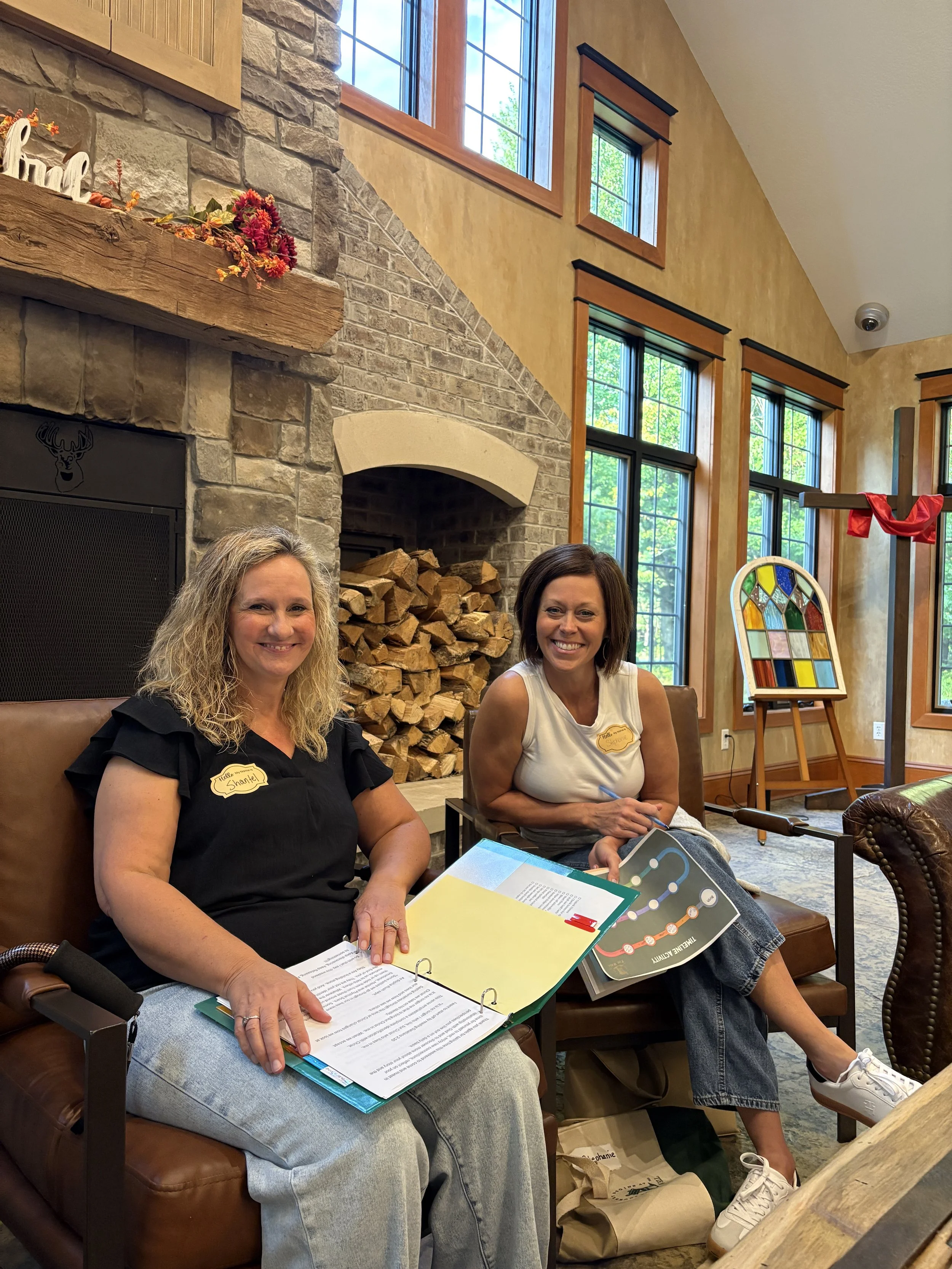 Two women sitting on chairs in a cozy, rustic living room with a stone fireplace and wood logs, large windows, a stained glass window, and a cross with a red cloth.