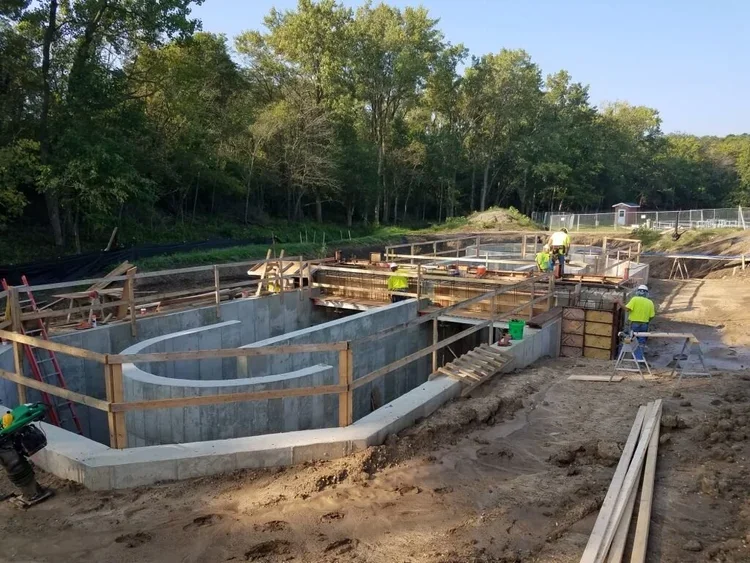 Construction workers with concrete walls and wooden supports, surrounded by trees and clear skies.