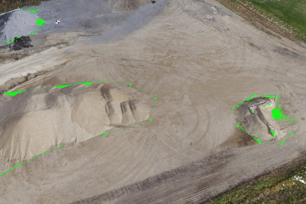 Aerial view of a construction site with two large excavated areas outlined in green, containing piles of sand or soil, and a gravel surface surrounding the excavations.