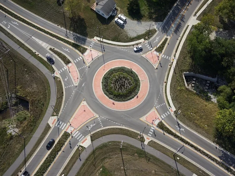 Aerial view of a roundabout with four roads converging, central island with greenery, and marked crosswalks and bike lanes.