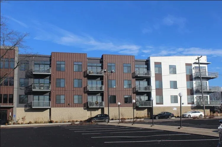 A modern apartment building with three floors, featuring brown and white panels, multiple balconies, and large windows, situated on a street with parked cars and streetlights under a clear blue sky.