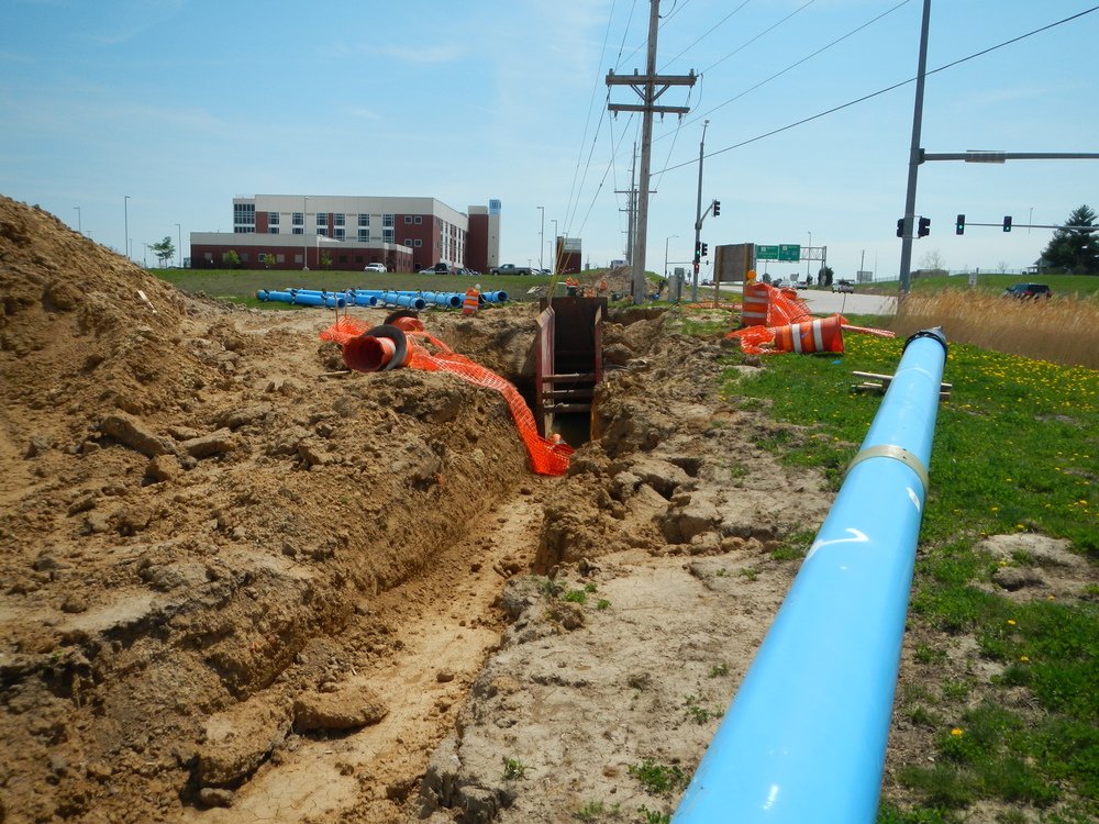 Construction site with large blue pipe, orange safety fencing, and excavated trench along roadside, near utility poles and a building in the background.