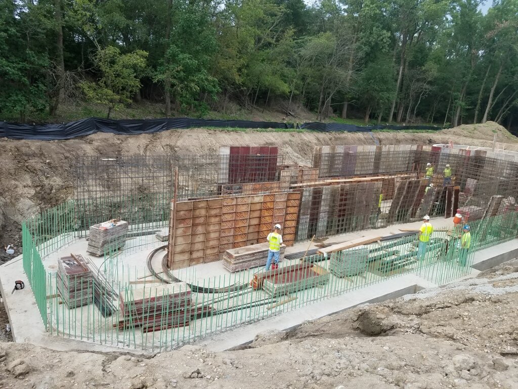 Construction workers in safety gear working on building foundation with steel rebar and wooden formwork in a wooded area.