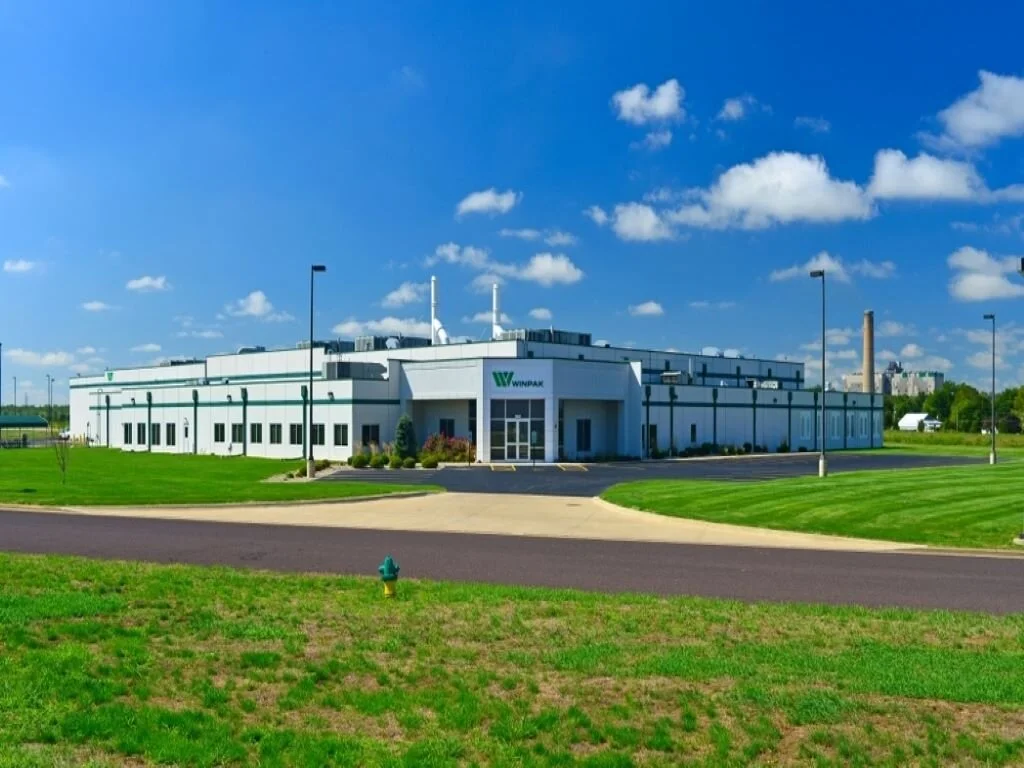 A large white industrial building with the sign 'Winpak' on it, surrounded by green lawns, a paved driveway, and a bright blue sky with some clouds.