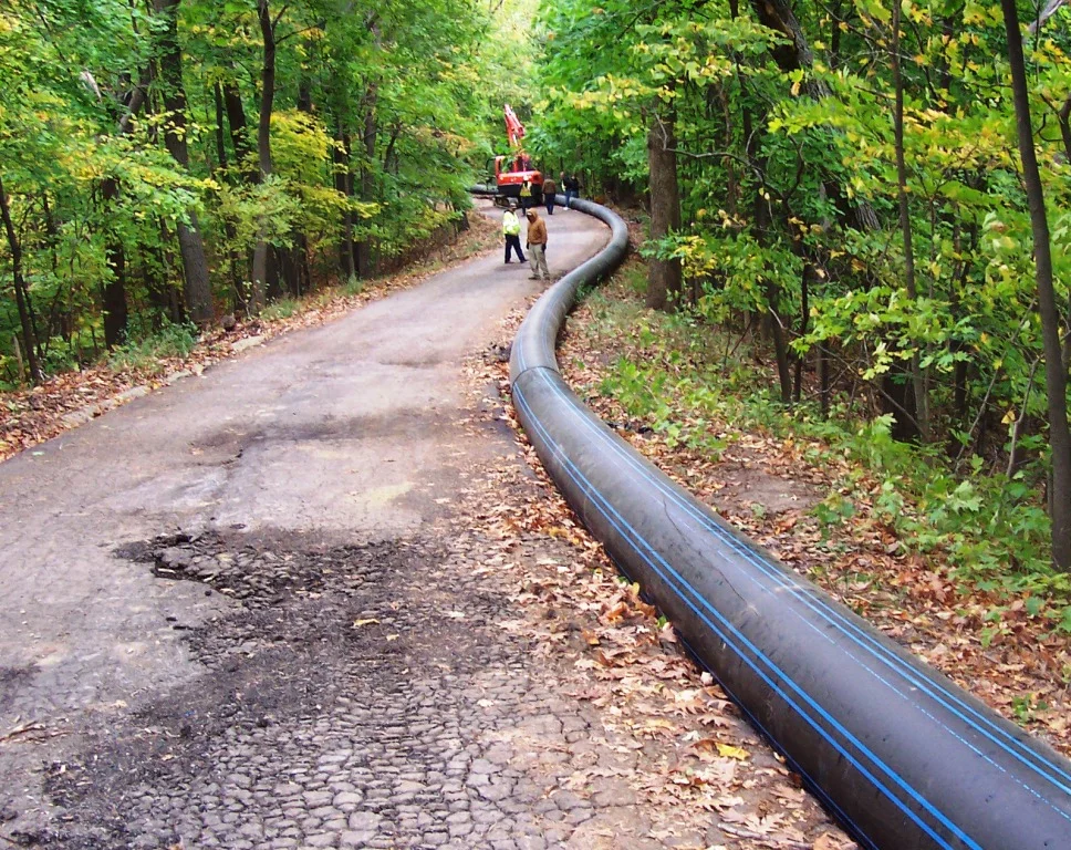 A large black pipeline with blue markings running along a dirt road through a green wooded area. There are workers and a truck in the distance near the pipeline.