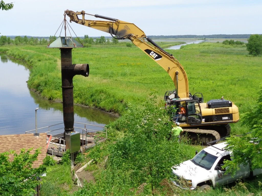 A construction excavator lifting and installing a large pipe in a waterway near greenery.