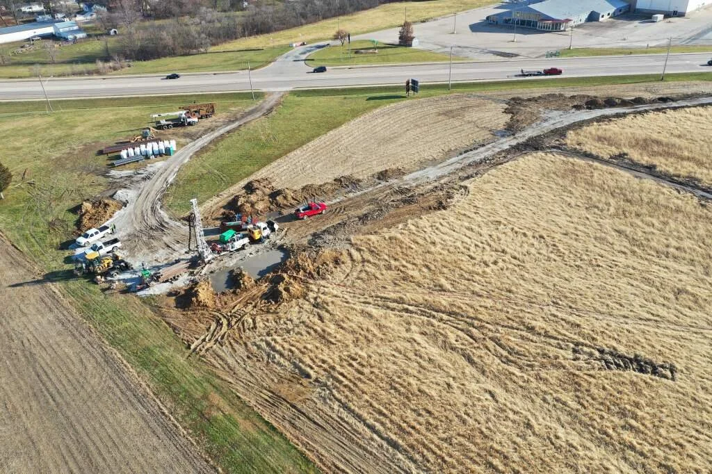 Construction site with machinery and vehicles on a patch of disturbed earth near fields and a highway.