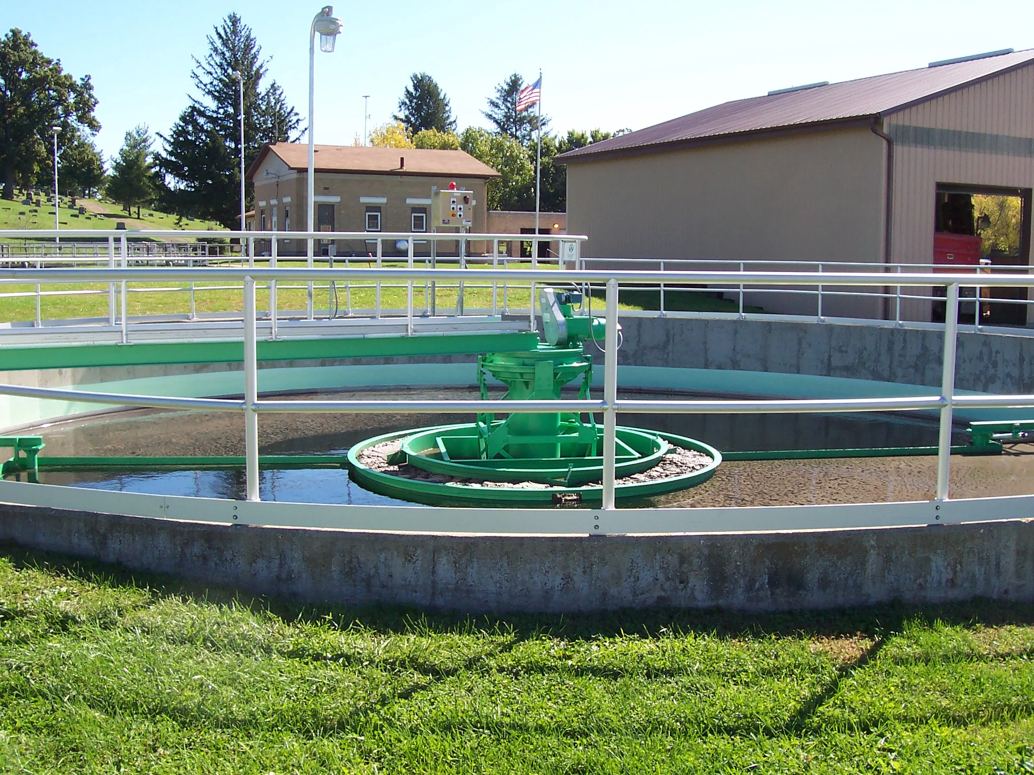 A circular clarifier tank with handrails and green mechanical components used in water treatment, situated outdoors on a grassy area with buildings and trees in the background.