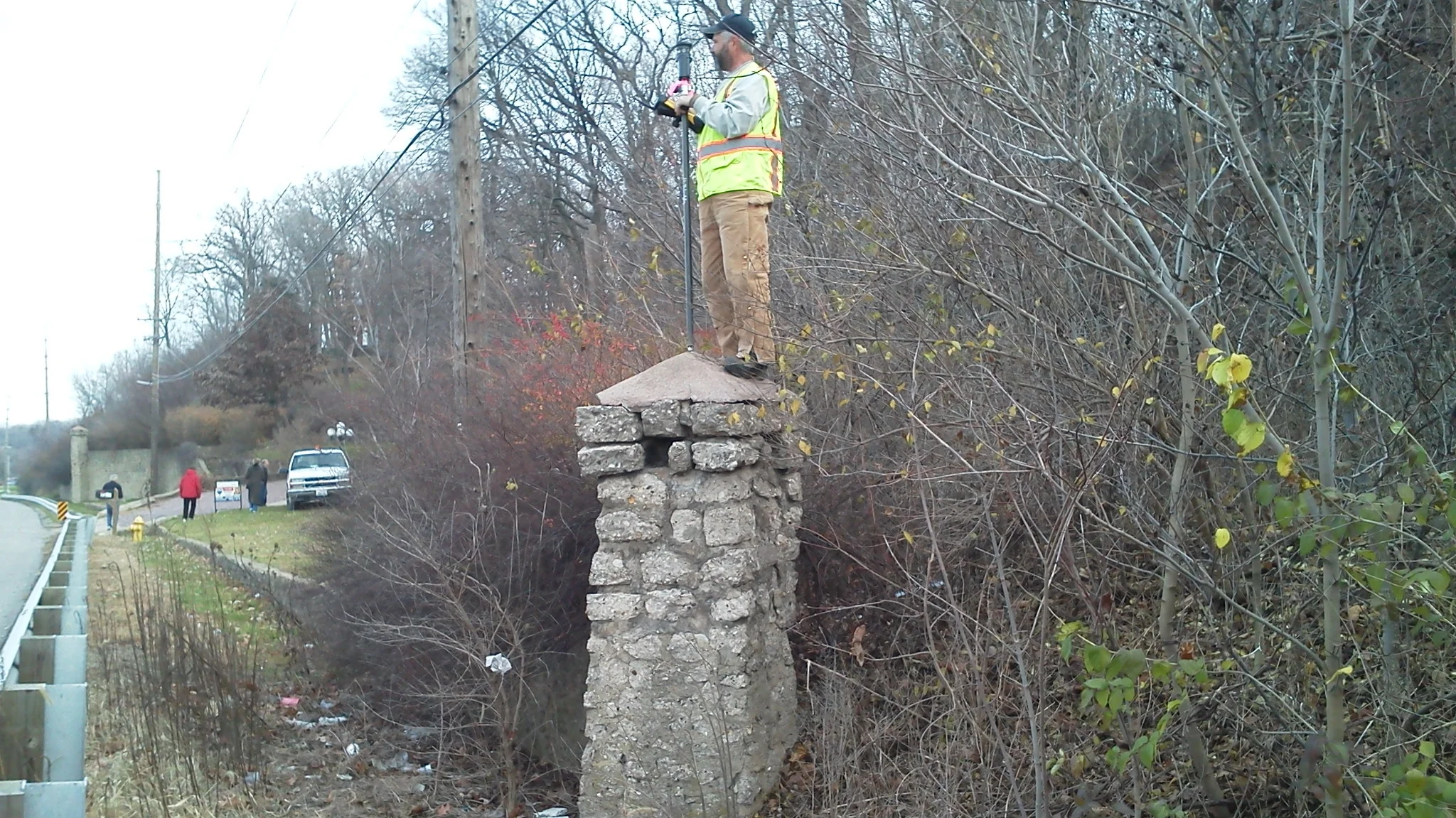 A worker with a safety vest and helmet standing on top of a stone pillar, using a tool to survey.