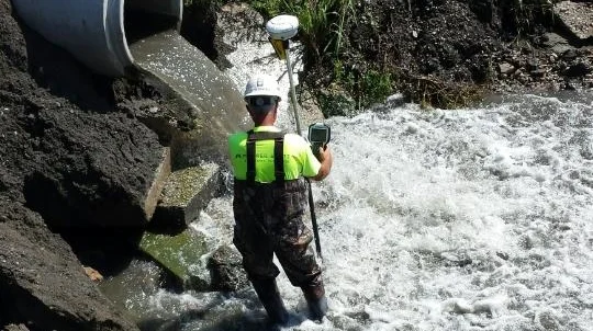 A worker wearing safety gear, including a helmet and reflective vest, standing in fast-moving water.