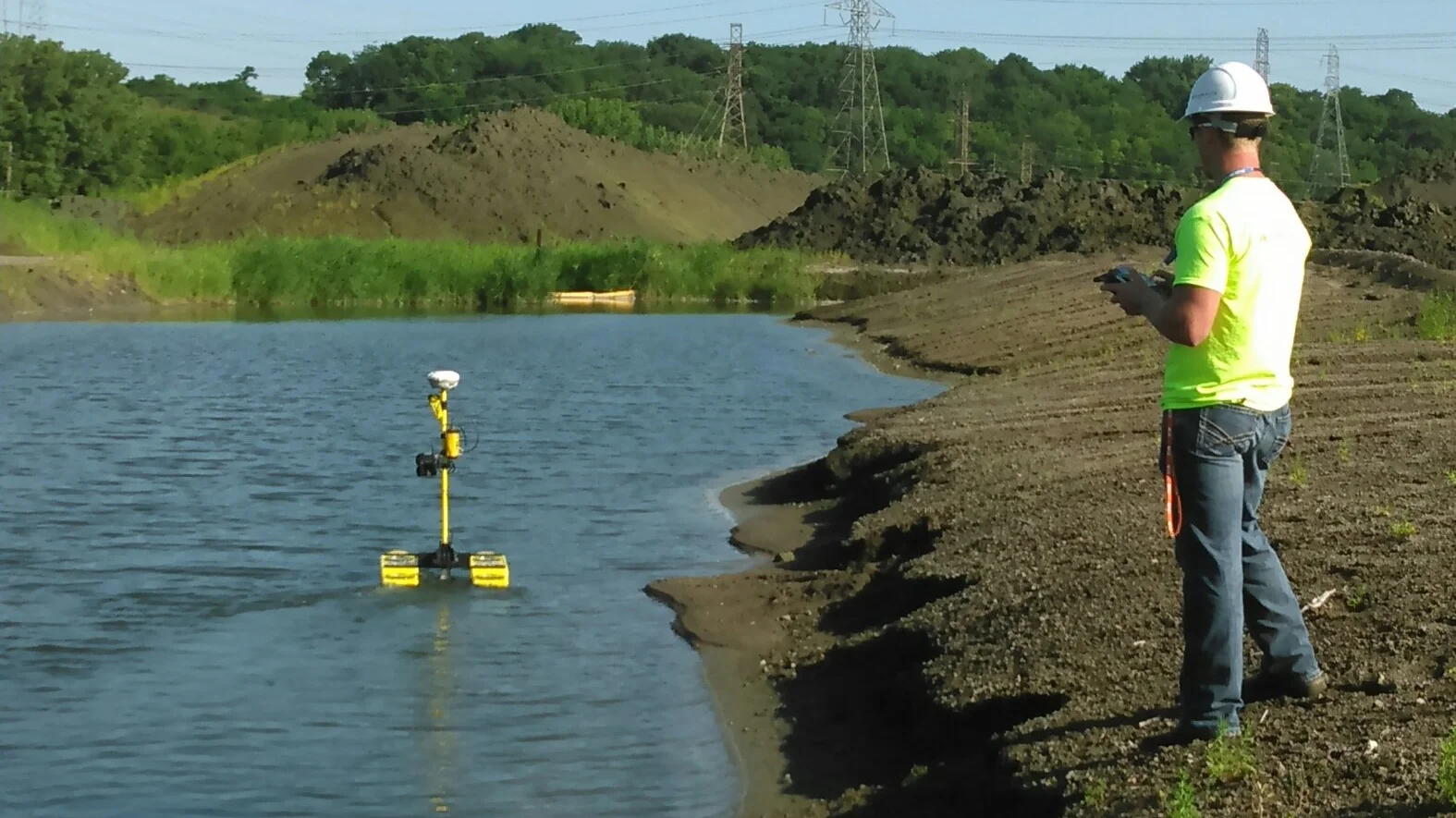 A worker in a safety helmet and neon yellow shirt operating a remote control on a dirt shoreline near a body of water, with surveying equipment floating in the water.