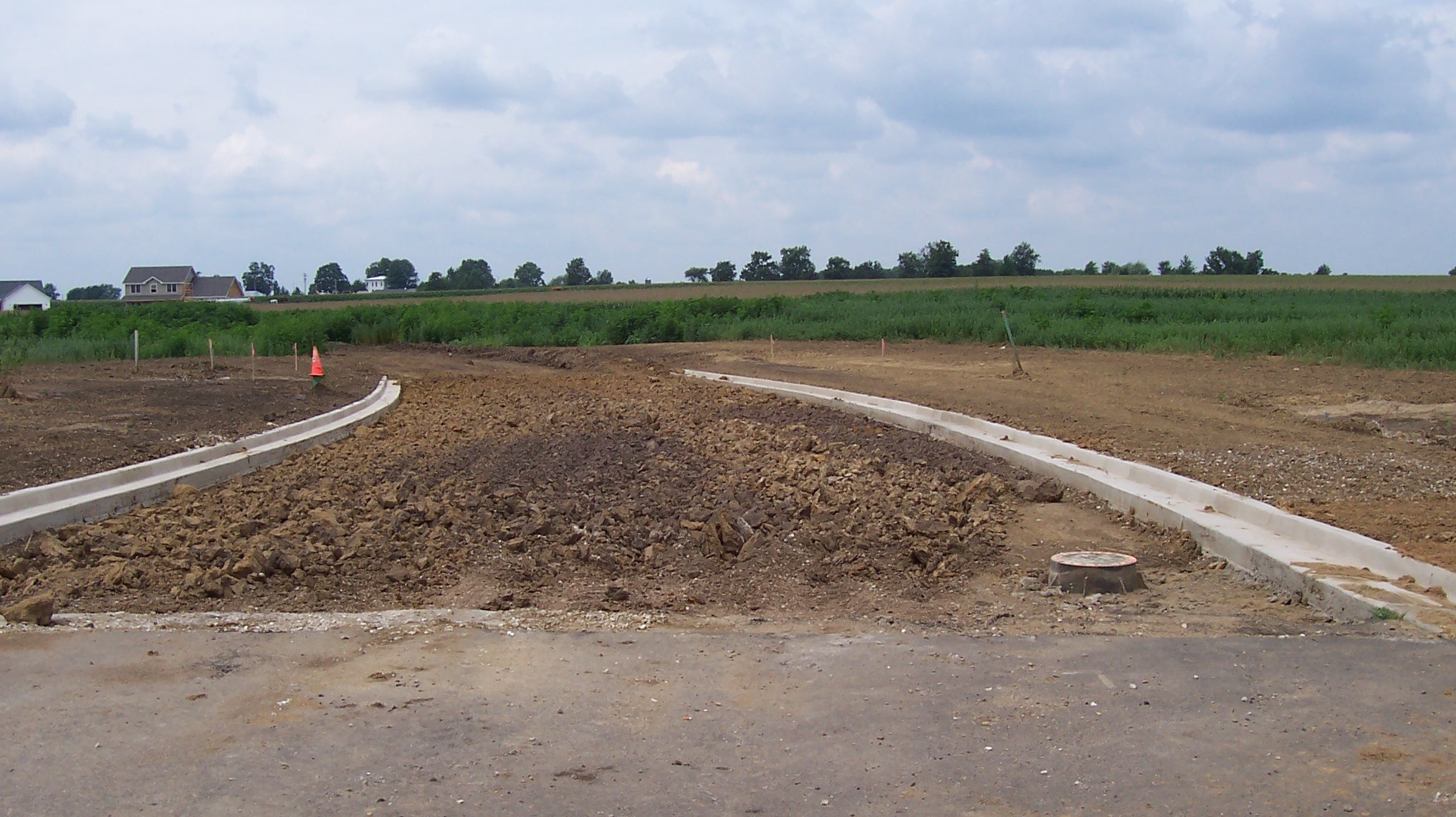 Construction site with a partially completed road, dirt and gravel, and orange safety cones under a cloudy sky