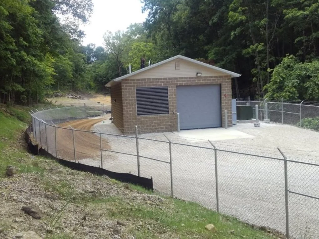 A small building with a brick lower wall, a gray door, and a vent, located within a fenced area on a dirt road in a forested area.