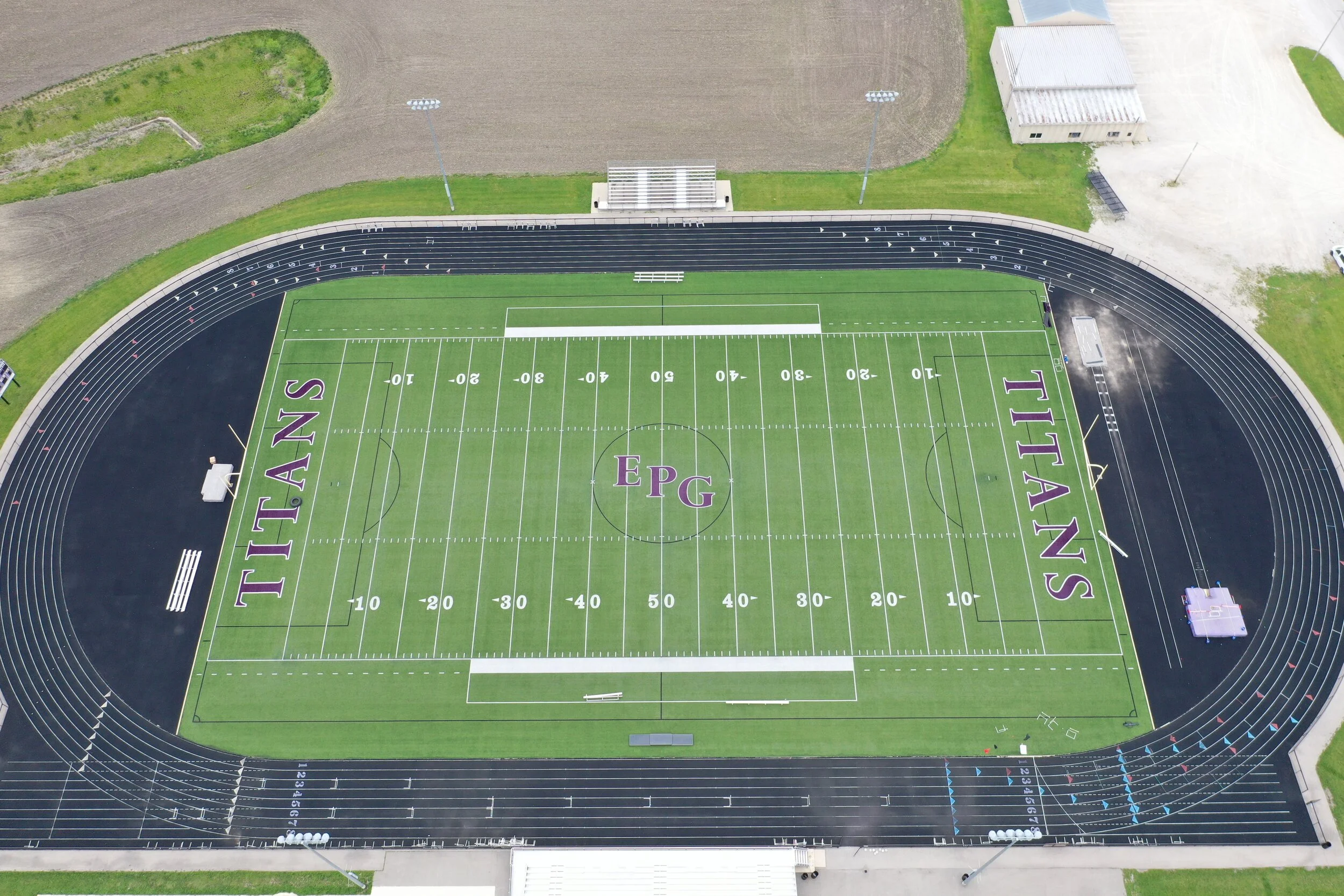 An aerial view of a football field with the word "TITANS" written on each end zone, the center circle marked with the letters "EPG," and the initials "TITANS" on the sideline. The field is surrounded by a black running track, with some outdoor lights and a small building nearby.