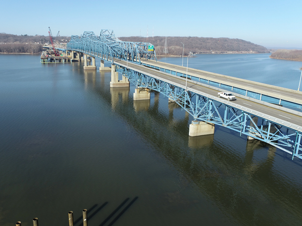 A blue steel truss bridge over a large body of water, with a few cars traveling across it and construction equipment visible on the far end.