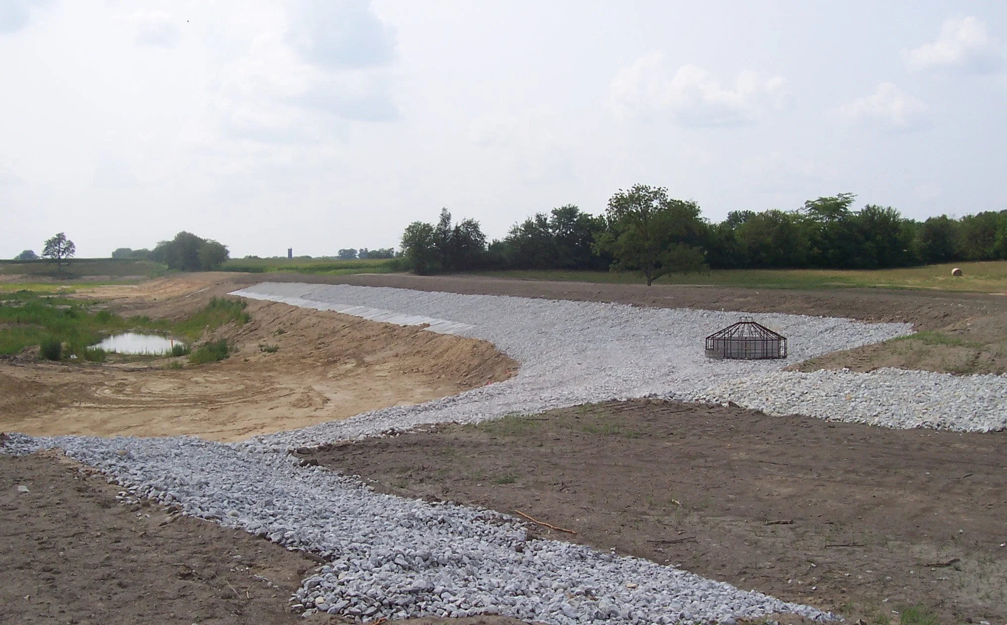 A landscape with a winding gravel path, a small pond, trees, and a greenhouse frame under a partly cloudy sky.