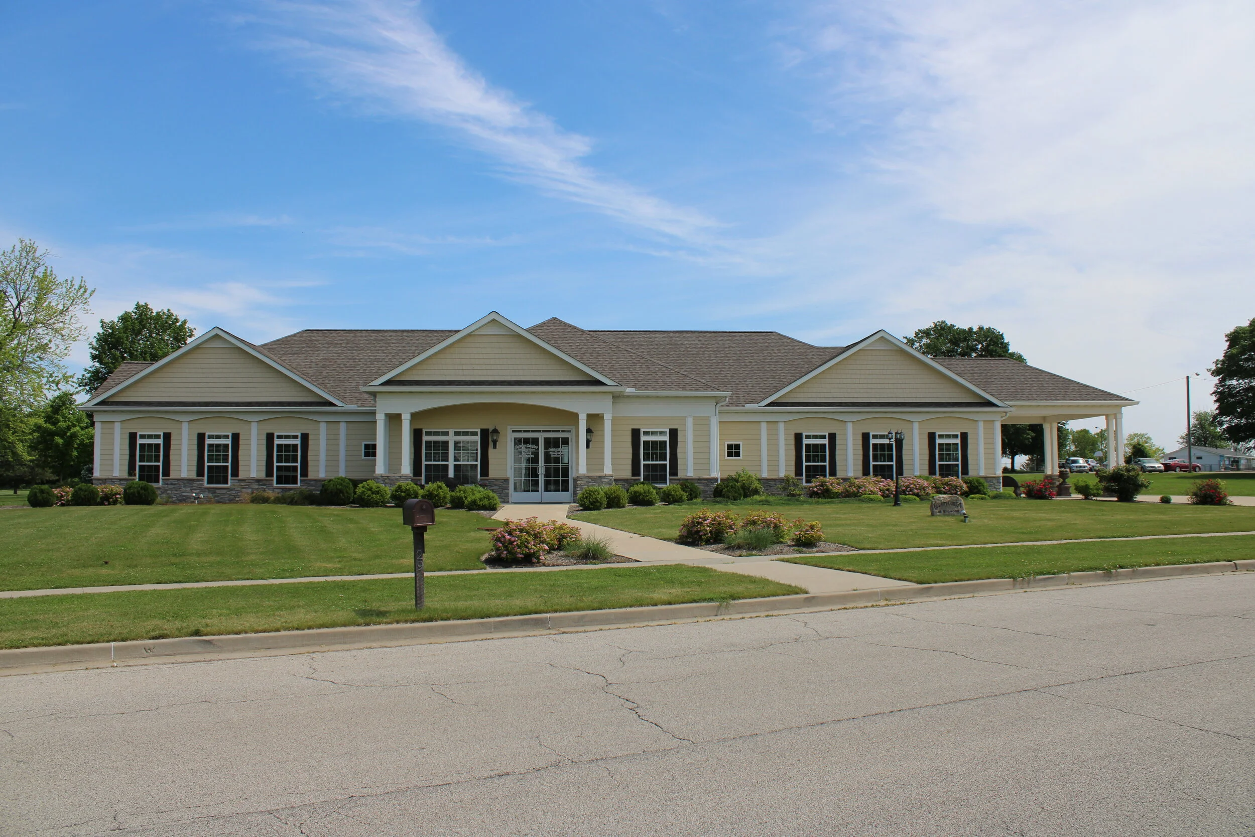 A large, single-story yellow house with white trim, a stone foundation, and multiple windows, surrounded by a well-kept lawn with bushes and flowers, on a sunny day with a blue sky.