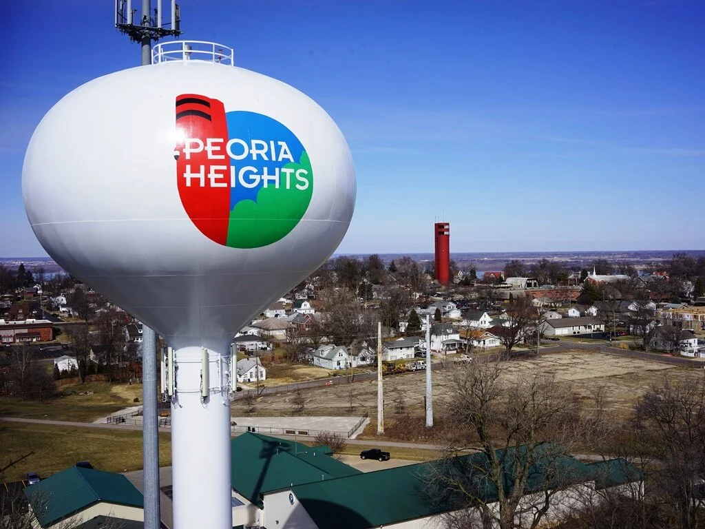 A water tower with a Peoria Heights logo over a residential neighborhood with trees, houses, and a distant red water tower under a blue sky.