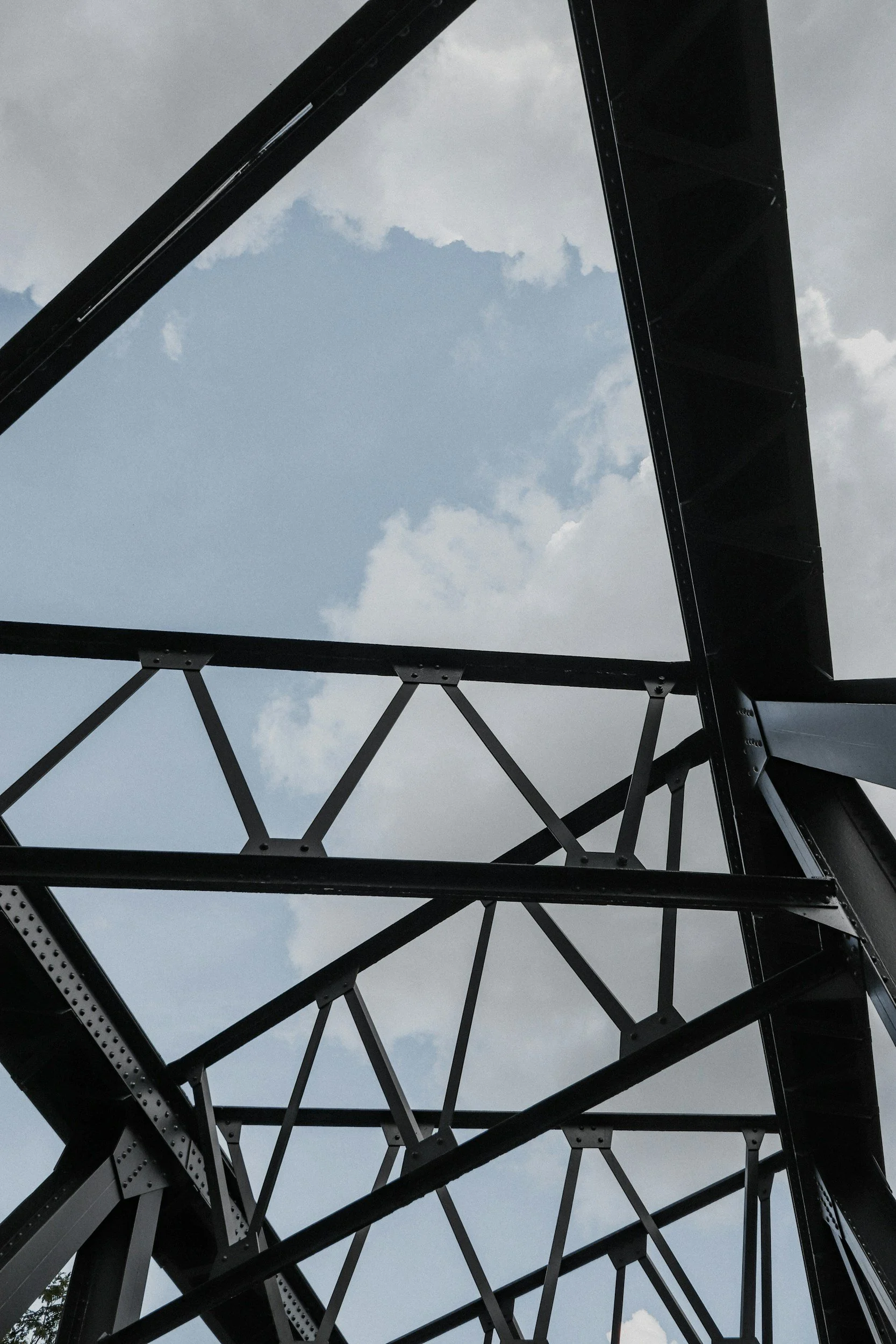 Looking up at a black steel bridge structure with geometric beams and trusses against a partly cloudy sky.