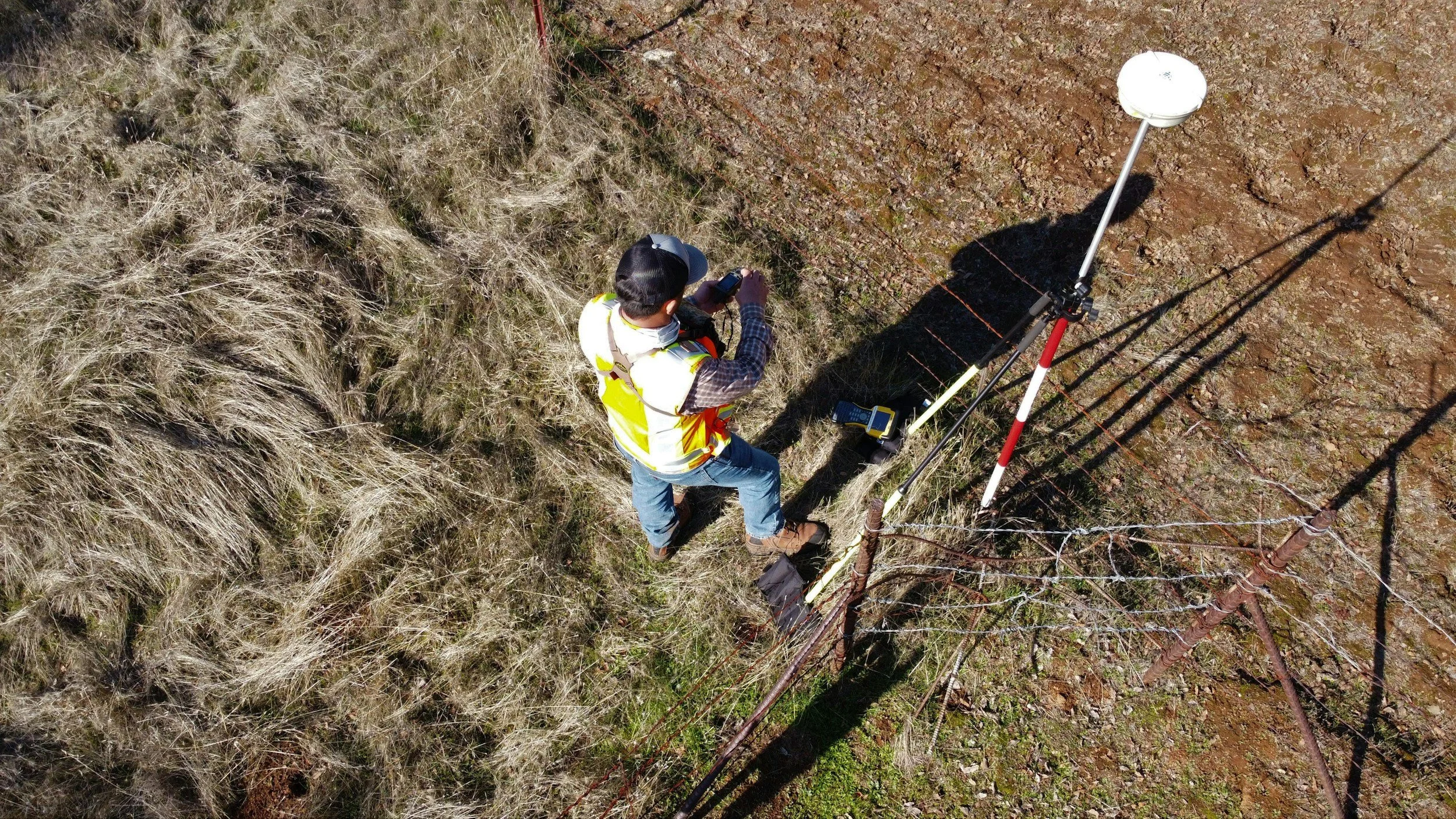 A person wearing a yellow safety vest and a cap working on a utility pole during daytime, using specialized equipment in a grassy and rocky area.