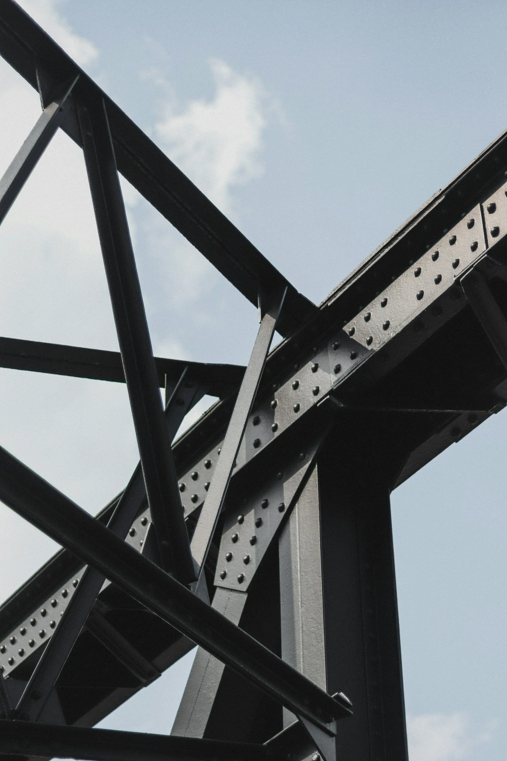 Close-up view of a black steel bridge truss structure against a blue sky with scattered white clouds.